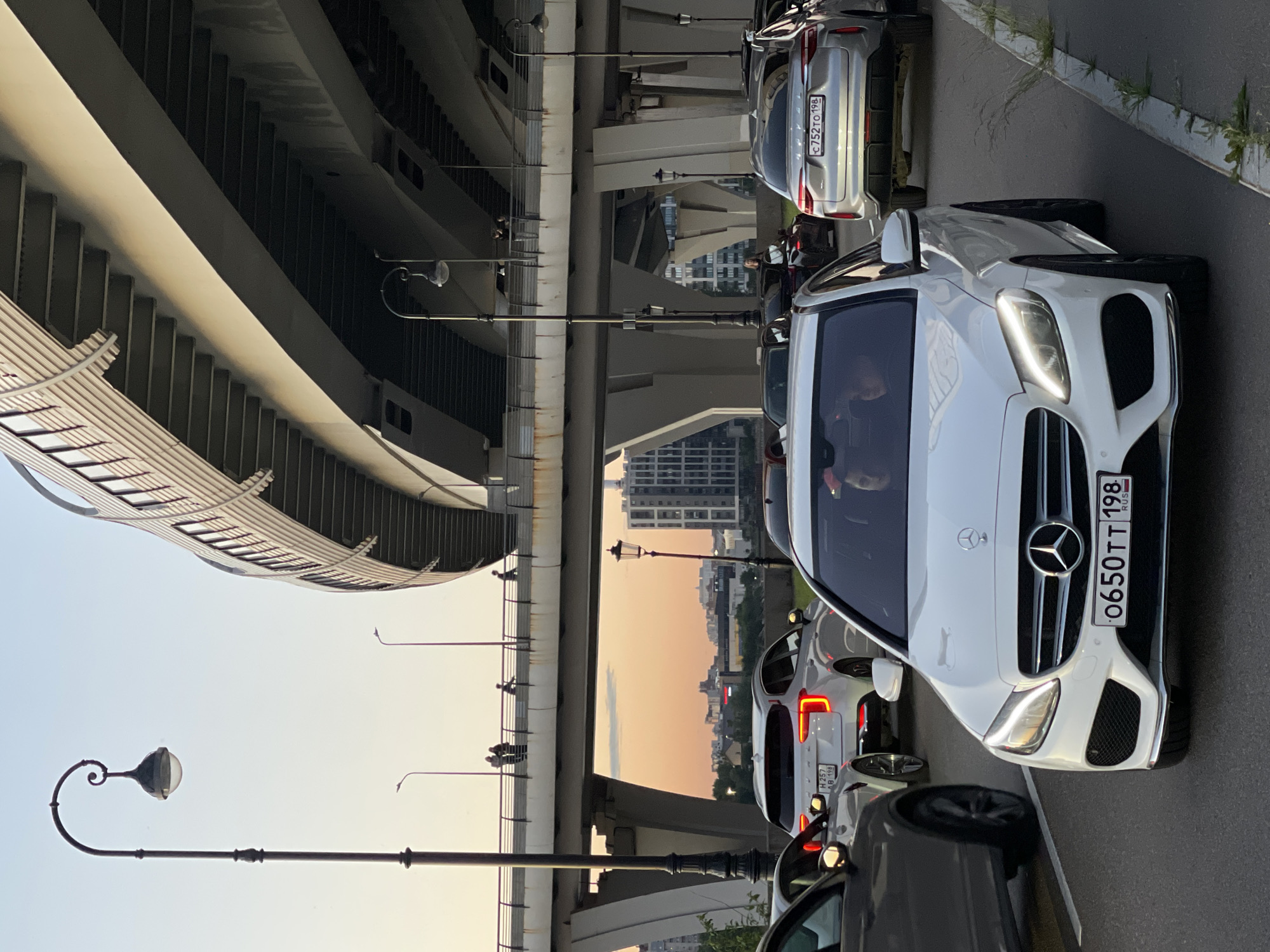 A wide shot captures a traffic jam on a street beneath a massive elevated highway or bridge structure in an urban environment. The sky is clear with a soft gradient of light, indicating either early morning or late afternoon. In the foreground, a white Mercedes-Benz C-Class is prominently featured, its headlights illuminated. Behind it, a silver SUV, possibly a Haval, is also visible. The highway structure above is a dominant feature, with multiple levels and supporting pillars creating a complex, layered effect. Several lampposts are present on the street level, some with decorative tops. In the background, city buildings and architecture are visible through the gaps in the highway structure, suggesting a dense urban landscape. A few pedestrians can be seen on an elevated walkway or pedestrian bridge connected to the highway structure, adding a human element to the scene. The overall impression is one of a busy, modern city with extensive infrastructure.