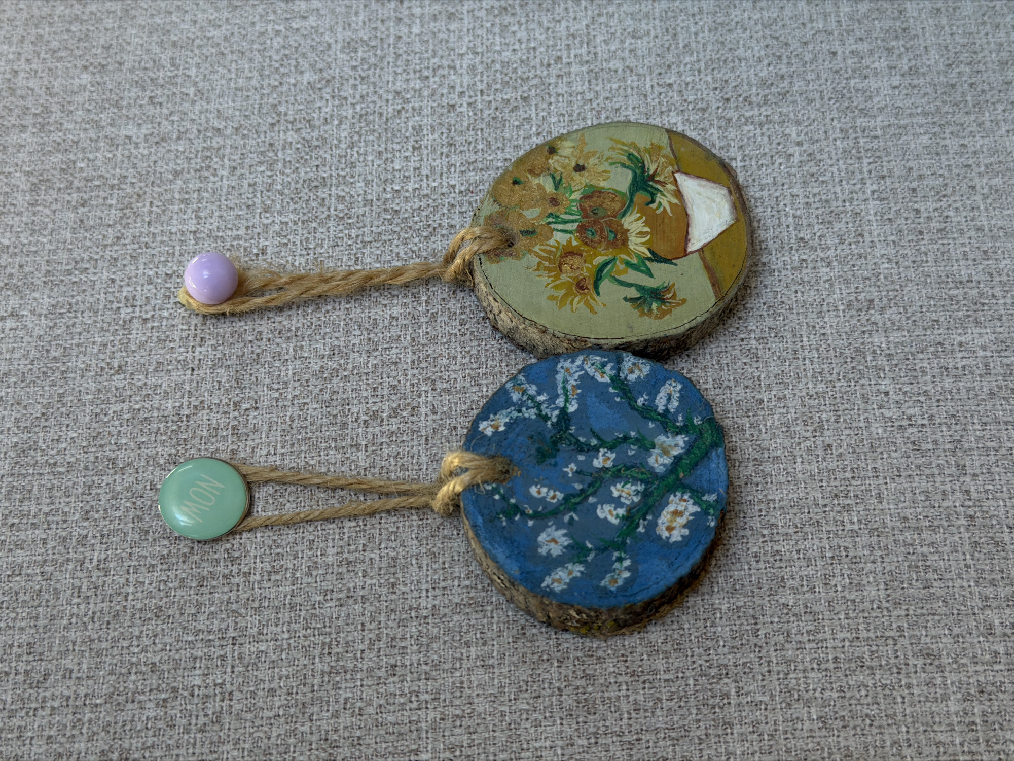 Two painted wooden disk ornaments lie on a gray textured surface. The top ornament depicts a still life of sunflowers and is attached to a piece of twine with a light purple bead on the end. The bottom ornament is painted with a blue background and white blossom-like flowers. This ornament also has a piece of twine tied to it, with a light green button that reads "MON" on the end. The items appear to be handmade crafts. There is no information in the image to suggest this is located in Yerevan, Armenia.