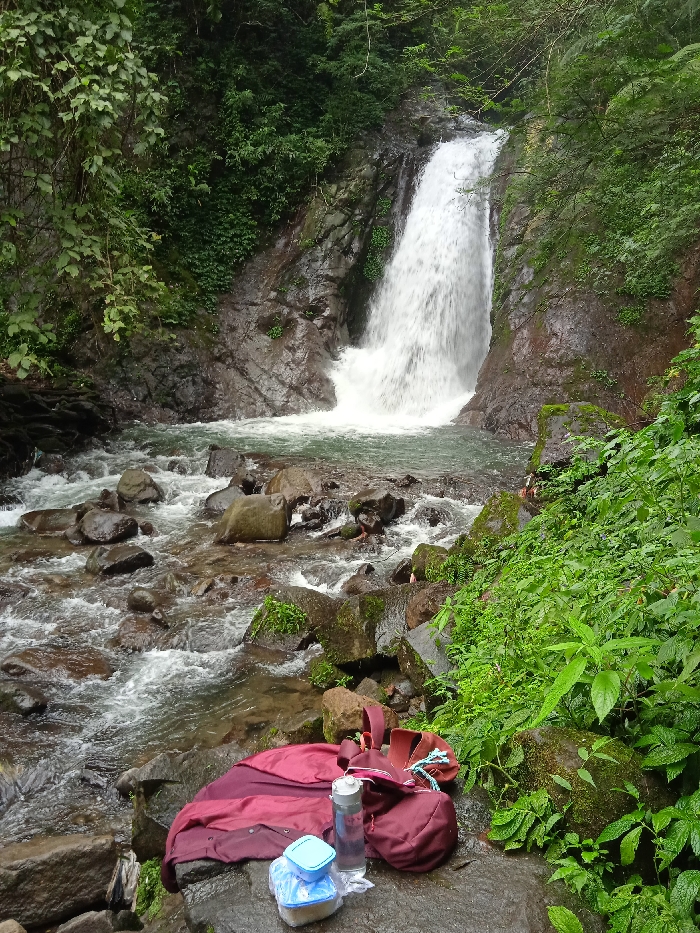 The image shows a waterfall in Tawangmangu, Indonesia, at the coordinates -7.655 latitude and 111.135 longitude. The waterfall cascades down a rocky cliff face, creating a pool of water at its base. The water flows through a riverbed with many rocks. Lush green foliage surrounds the waterfall. A red jacket, a backpack, a blue water bottle, and a blue container are placed on the rocks in the foreground. The water is clear and flowing rapidly. There are no people visible in the image. The scene suggests a peaceful and serene natural environment.