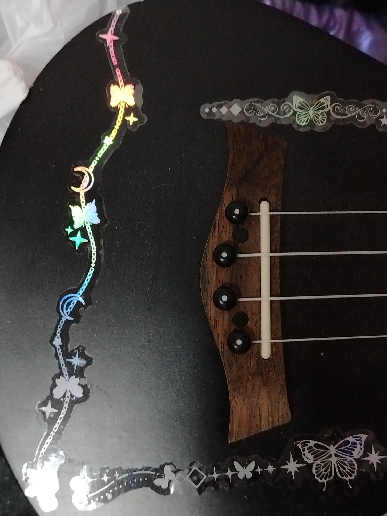 A close-up, angled shot features a black ukulele adorned with iridescent, holographic stickers. The dark surface of the instrument provides a striking background for the shimmering decals. Along the upper left curved edge, a decorative strip of stickers forms a whimsical, chain-like pattern, showcasing stars, crescent moons, and butterflies that display a rainbow gradient effect as light reflects off them. Another strip of stickers is visible along the top right edge, above the instrument's bridge, featuring intricate swirling patterns, diamond shapes, and a central butterfly, also with a subtle iridescent sheen. A third section of similar stickers, including a large butterfly, stars, and diamond shapes, is visible along the bottom right edge of the ukulele.
The ukulele's dark brown wooden bridge, with its visible grain, is centrally positioned. Four white nylon strings extend from the bridge, secured by four black bridge pins. No people are present, and the instrument appears to be stationary. The lighting highlights the reflective qualities of the stickers. The background is dark and out of focus, suggesting an indoor setting, possibly on a soft surface or within a case. No text is visible in the image. This scene takes place in Izhevsk, Russia.