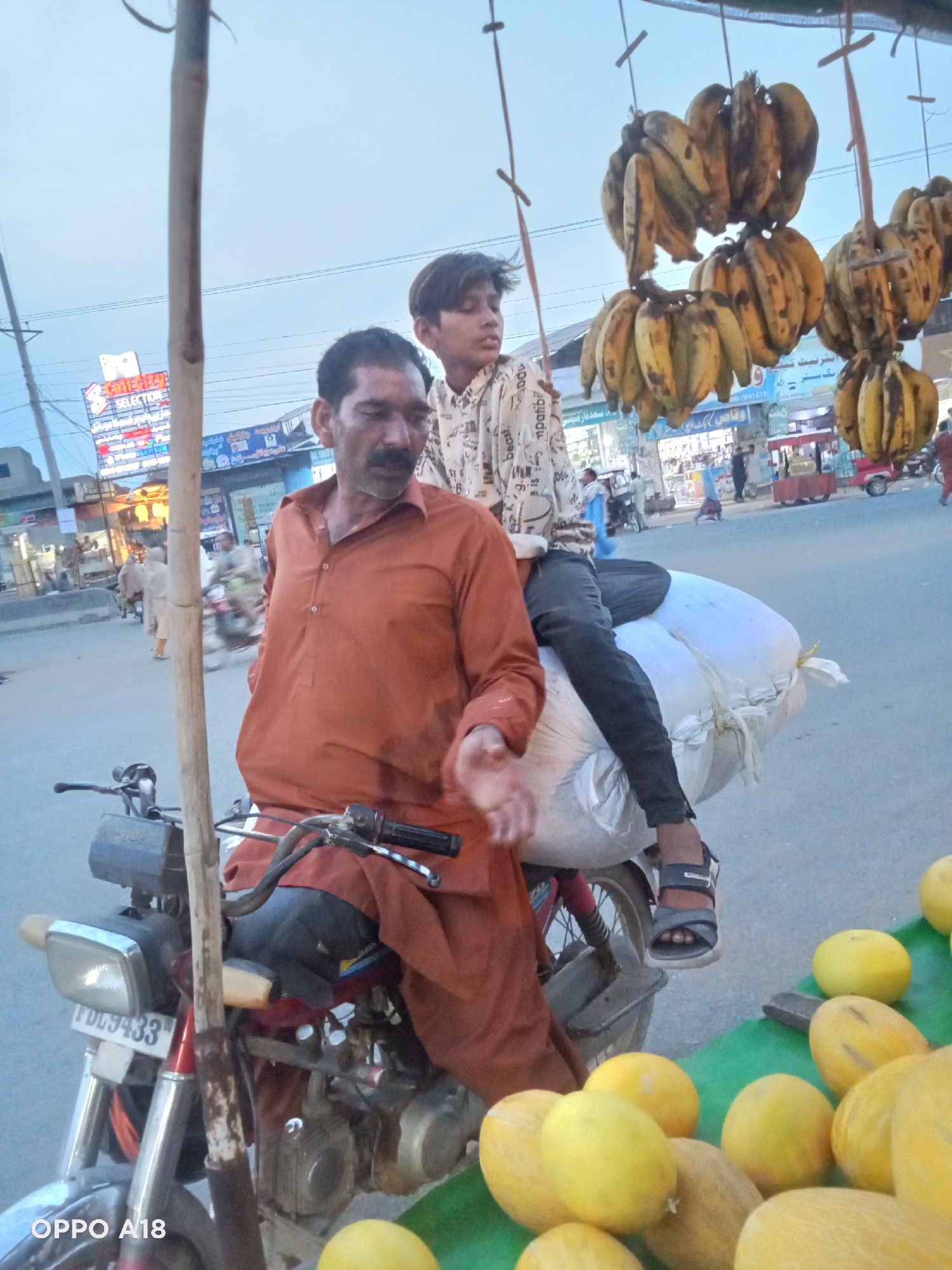 The image captures a vibrant street scene, likely in South Asia, centered around a roadside fruit vendor. In the foreground, an adult man with a mustache, dressed in an orange shalwar kameez, sits on a motorcycle. His right hand is outstretched, as if gesturing or presenting something. Behind him, a younger boy sits atop two large white sacks strapped to the back of the motorcycle, looking to the side.
To the right of the man and boy, several bunches of bananas hang from a simple stand, indicating a fruit stall. At the bottom right, a collection of yellow melons are displayed on a green surface, further confirming the vendor's trade.
The background reveals a bustling street with various shops and buildings. Several other people are visible walking or riding motorcycles. The general lighting suggests it is either late afternoon or early evening, with a clear, light blue sky. The atmosphere appears busy, characteristic of a market street.
Notable text includes "PDG9433" on the motorcycle's license plate and "OPPO A18" as a watermark at the bottom left. In the background, shop signs display words like "SELECTION", "ASUM", "AL SHIPA", "beast.", "mpatibi", and a phone number "100-7081275". Some visible Urdu or Arabic script includes "نترنيت كيف" (internet how/quality) and "رنگی" (colorful).