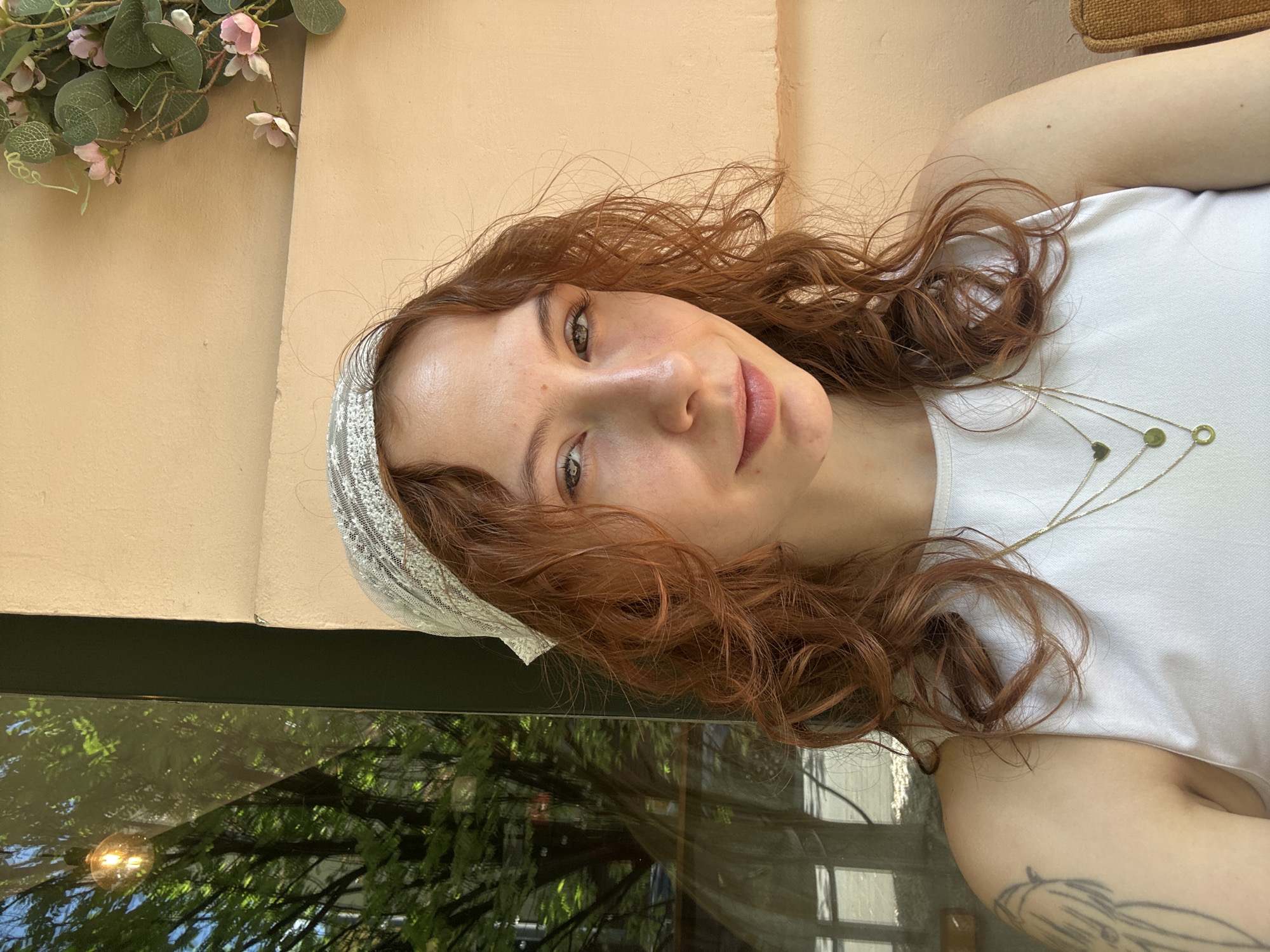 A young woman with reddish-brown, wavy hair and green eyes looks directly at the camera with a soft smile in this vertical portrait. She is wearing a white sleeveless top, a gold necklace with multiple small charms (including green and gold circles and a heart), and a light-colored, patterned headband partially obscured by her hair. A faint tattoo is visible on her left arm, near the shoulder.
The background consists of a light peach-beige colored wall. In the upper left corner, an artificial floral arrangement with pink blossoms and green foliage hangs on the wall. Below the woman, a dark, reflective surface, likely a glass table or window, mirrors a scene of vibrant green trees, a bright light bulb, and the outlines of distant buildings, suggesting an outdoor view or a space with large windows.
The image is well-lit by natural light, indicating it was taken during the daytime. The overall mood is serene and pleasant. The setting appears to be a cafe or a similar establishment with an outdoor or well-lit interior, located in Novi Sad, Serbia. No text is visible in the image.