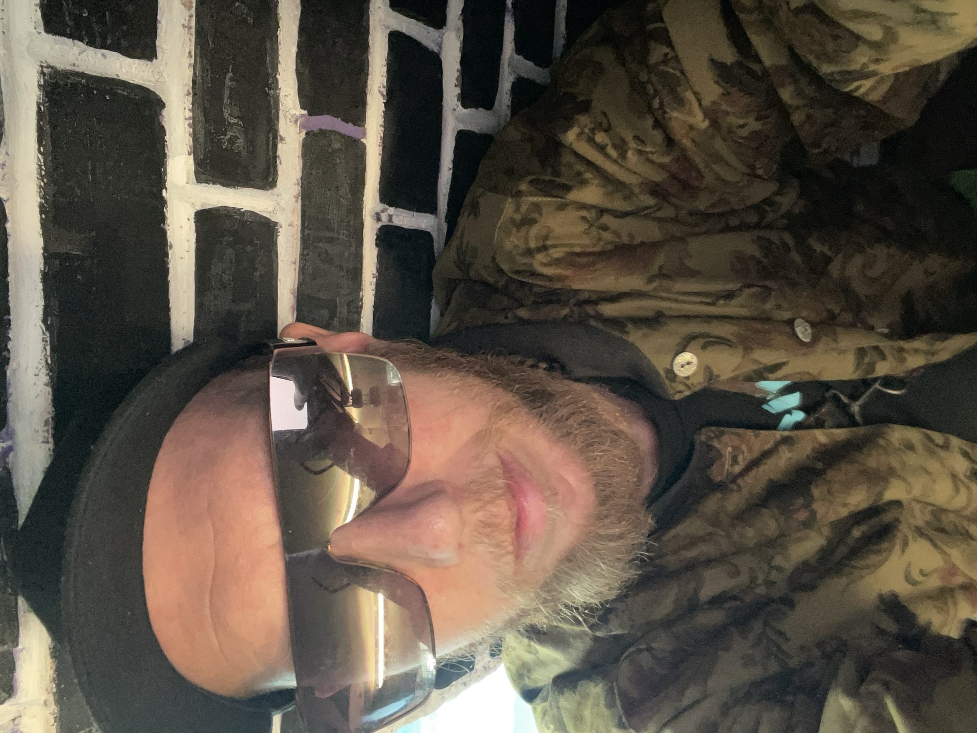 A close-up, overhead selfie shows a person wearing a black hat, large reflective sunglasses, and a camouflage jacket over a dark t-shirt. The person has a light complexion, a ginger beard, and is looking downwards towards the camera. The background consists of a black brick wall with white mortar, some of which has purple paint streaks. The overall lighting is dim, with reflections visible in the sunglasses. The image does not provide any information that would allow for the determination of the location within the city of Saint Petersburg, Russia.