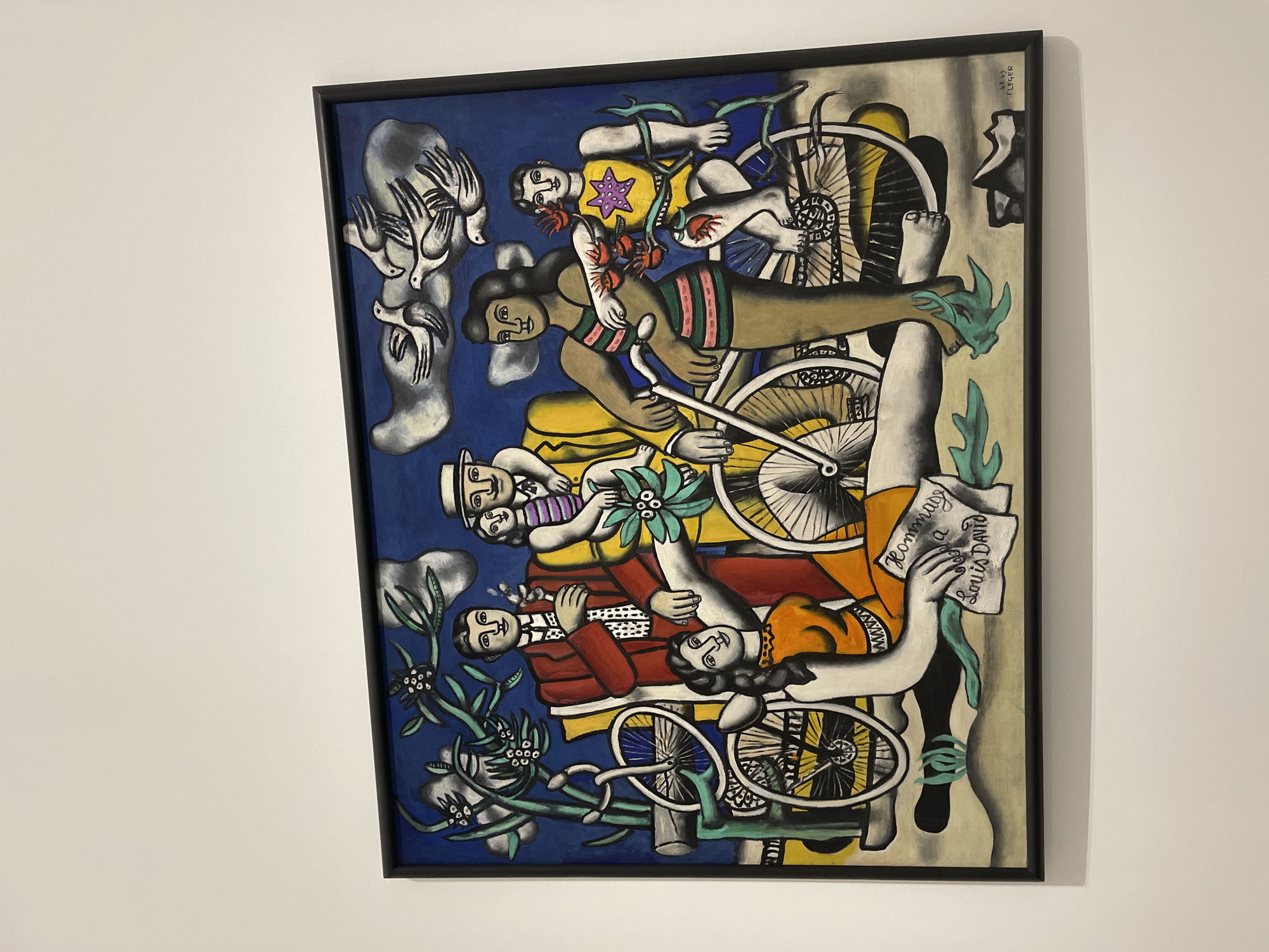 The image shows a framed painting hanging on a white wall. The painting depicts a surreal composition with figures, bicycles, and abstract shapes in a cubist style. The color palette is varied, including blues, yellows, oranges, reds, greens, and browns. The figures appear to be intertwined with the bicycles, and there are also elements such as birds and flowers. The inscription "Hommage a Louis David" is visible on a sheet of paper held by one of the figures. The painting is located in Metz, France.