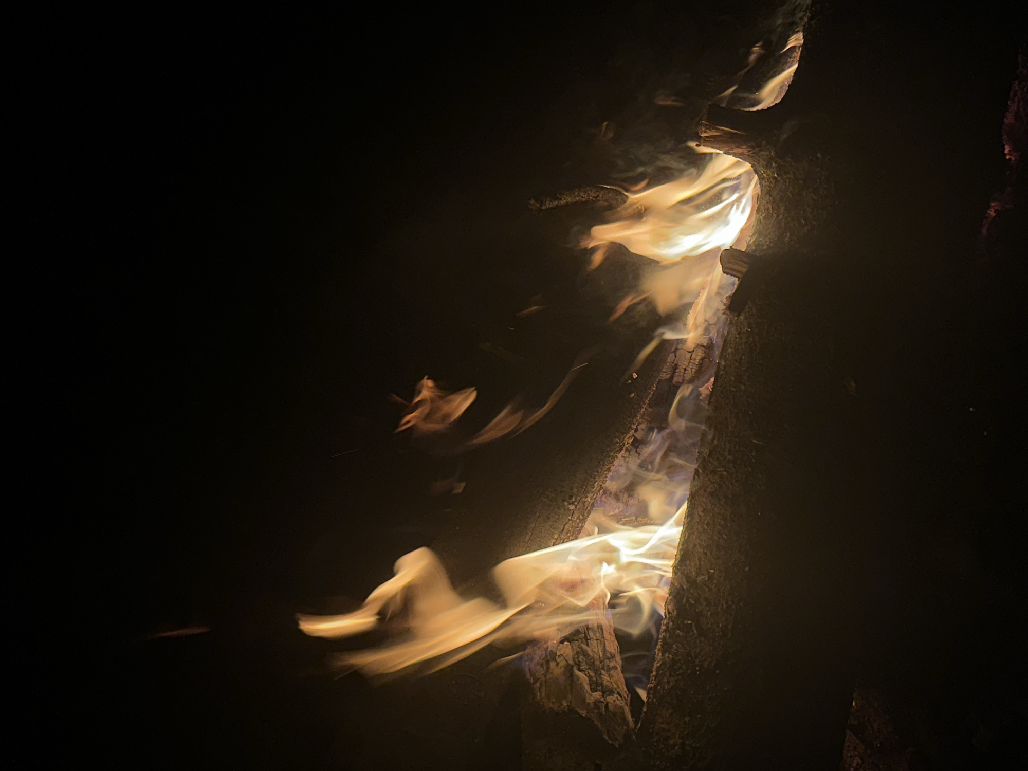 A vertical, close-up shot shows fire burning between two logs in the dark. The flames are yellow and white, with some blue hues. Smoke is visible. The logs appear rough and textured. The location is not evident from the image.