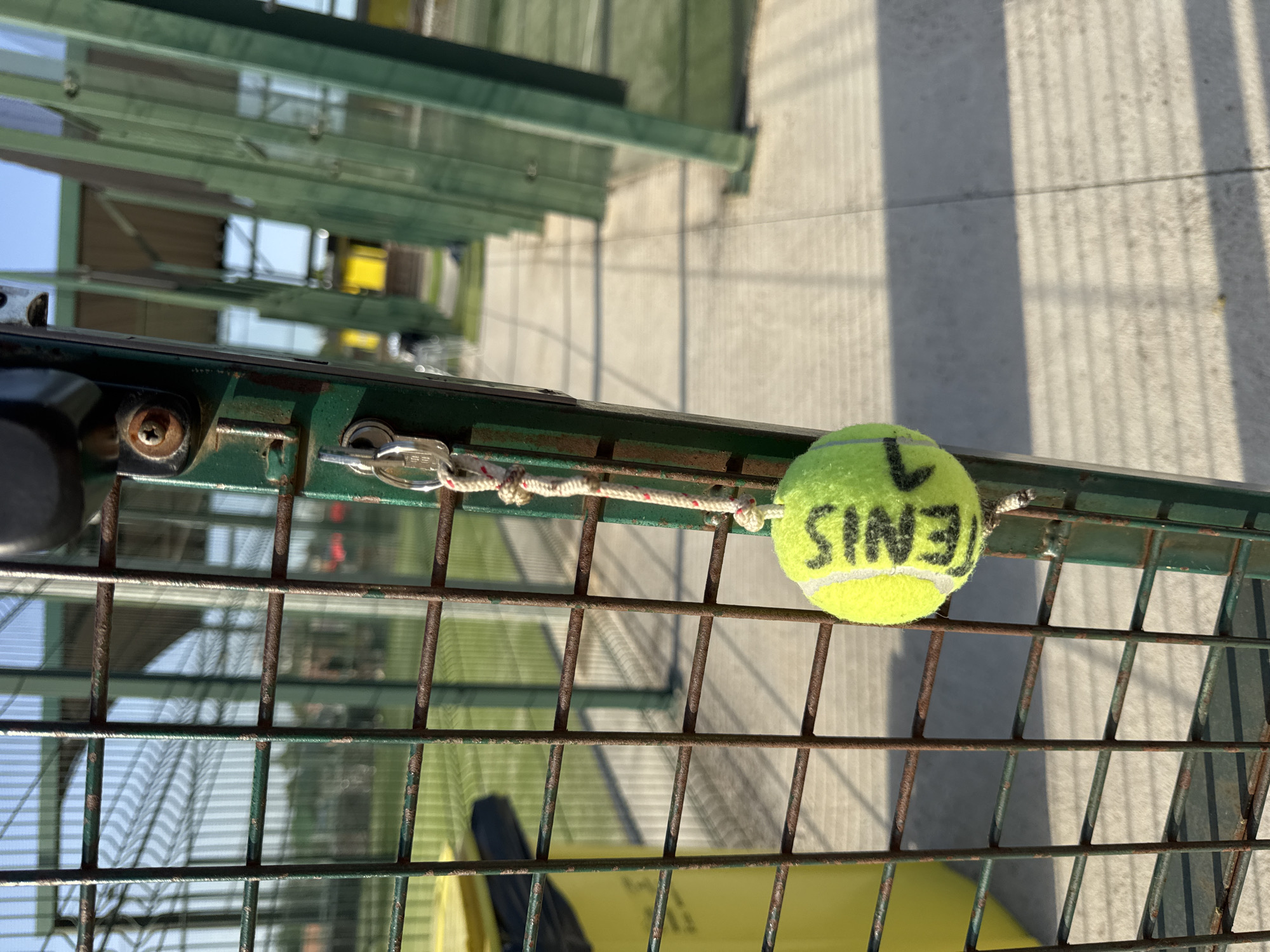 The image shows a close-up of a green metal fence with rusty bars. Hanging from the fence by a rope and a key ring is a tennis ball. The tennis ball is lime green with black writing on it, which appears to read "TENIS." The background is out of focus, but it appears to be an outdoor sports facility, possibly a tennis or paddle tennis court, with green fencing and a paved walkway. The sky is clear and blue. The overall lighting suggests it is daytime, likely late afternoon given the long shadows cast by the fence. No people are visible in the image. The scene is likely located in Vidreres, Spain, as indicated by the prompt, suggesting this is a public or private recreational area within that municipality.