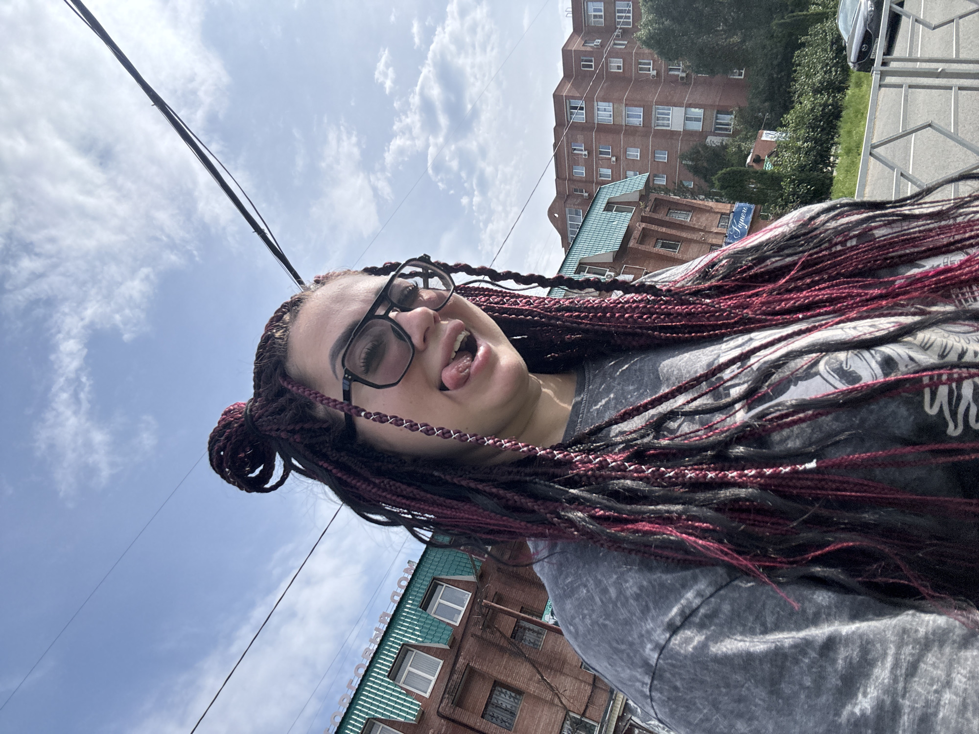 A young woman with braided hair, some of which is dyed a dark red, is taking a selfie outdoors. She is wearing glasses and a grey t-shirt. Her tongue is sticking out, and she is wearing large, black-rimmed glasses. In the background, there are brick buildings, trees, and a street with a car. The sky is blue with some clouds. The building in the background appears to be a residential building, likely in Tolyatti, Russia, given the Cyrillic text on the green awning of the building on the left, which reads "КОРПОРАТИВНЫЙ ДОМ" (KORPORATIVNYY DOM), meaning "Corporate House" or "Company Building". The overall environment suggests an urban setting on a partly cloudy day.
