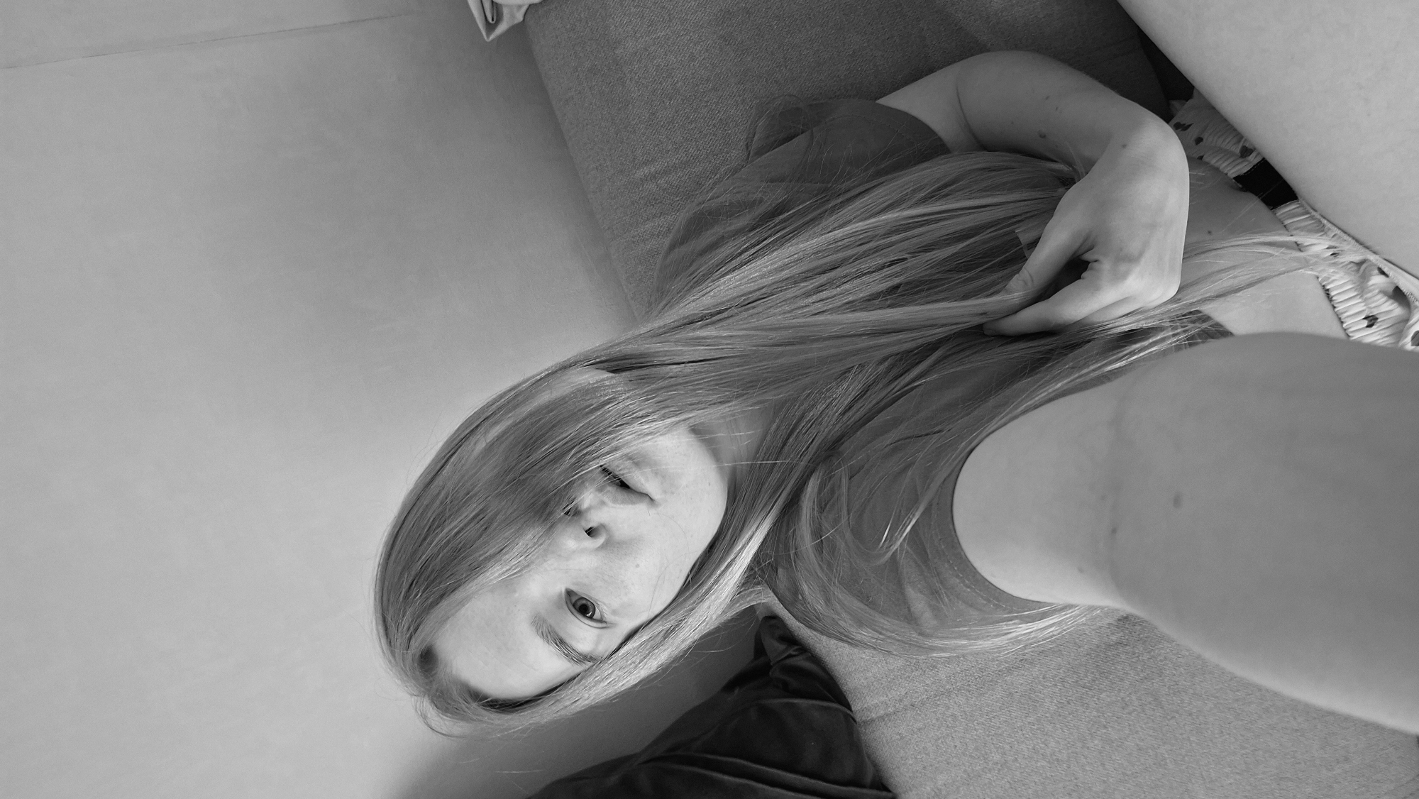 A black and white, close-up shot shows a woman lying down. The woman has long, light-colored hair that partially covers her face. Her hand is resting on her hair. She appears to be resting on a gray, upholstered surface, possibly a couch. To the side is a plain wall, and a black pillow. The location is unspecified, and there is no indication of the city of Chaikovsky, Russia.