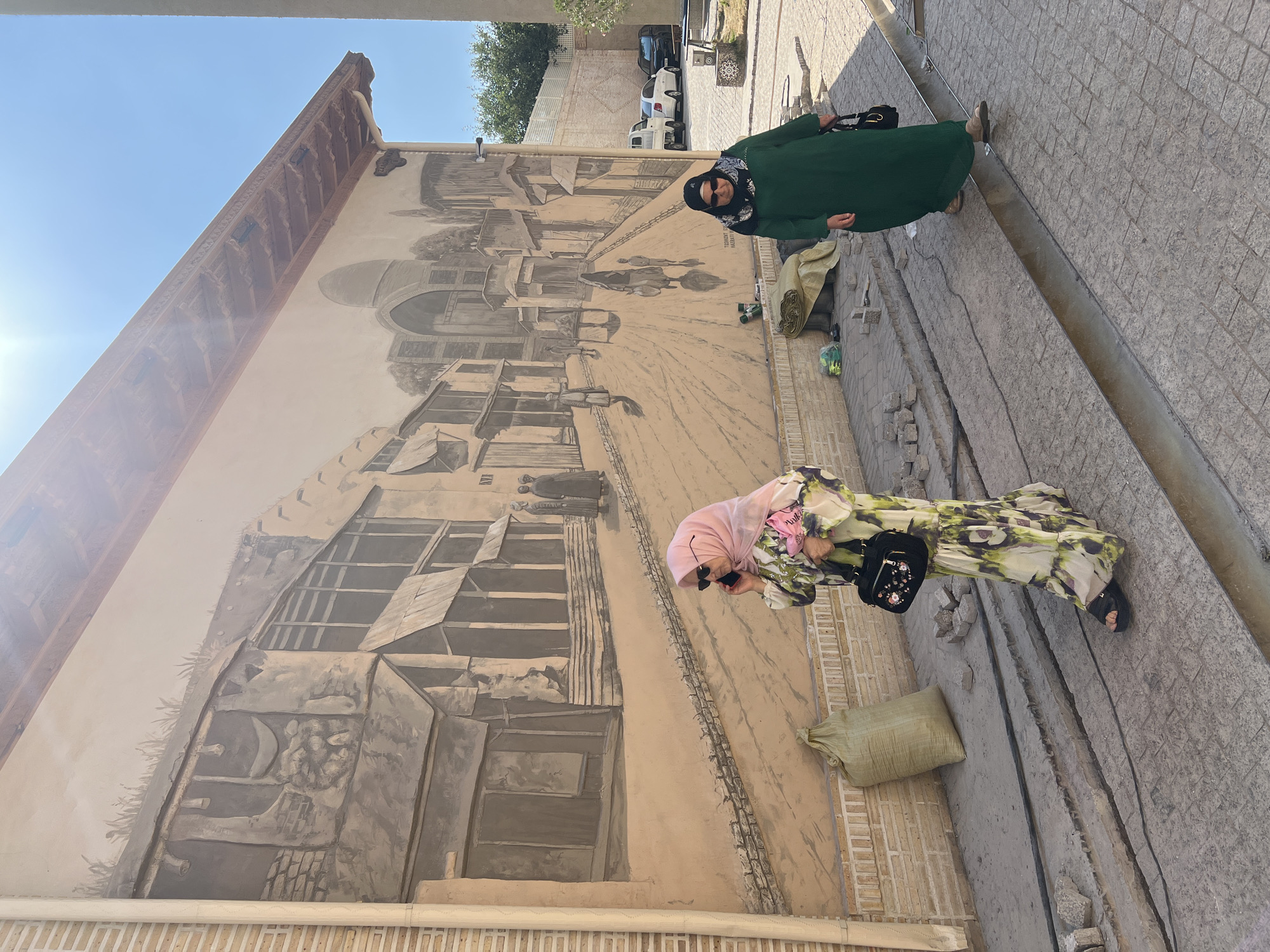 Two women are standing in front of a large mural painted on a building wall. The mural depicts a historical street scene with buildings, people, and carts. The woman on the left is wearing a green abaya and a black and white headscarf, with sunglasses. The woman on the right is wearing a long, floral-patterned dress and a pink headscarf, and is holding a black purse. Both women are looking towards the viewer, with mild smiles.
The scene is outdoors, with a clear blue sky visible at the top of the image. The ground is paved with cobblestones, and there are some sacks and building materials visible near the wall. The mural itself appears to be in grayscale and has a textured, bas-relief quality. Some of the mural depicts architectural elements such as arches and domes. There is some visible text on the mural, which appears to be in Arabic script.
The overall impression is of a sunny day in a historic location. The women appear to be tourists or residents enjoying the art. The mural is large and dominates the background, suggesting it is a significant piece of public art.