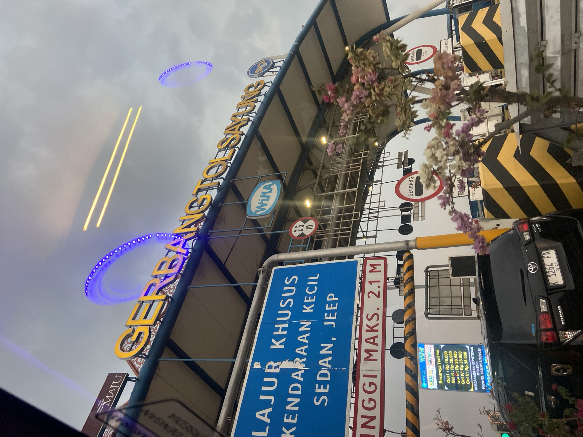 The image shows a toll gate in Sayung, Indonesia, identified by the prominent sign "GERBANG TOL SAYUNG" in yellow lettering. The toll gate features illuminated blue accents forming arches and circles in the sky. Several signs are visible: a blue sign indicating a lane for "KENDARAAN KECIL SEDAN, JEEP" (small vehicles, sedans, jeeps), and a red and white sign stating "TINGGI MAKS. 2.1 M" (maximum height 2.1 meters). There is also a circular sign with "WIKA" logo, another circular sign with a red border and black bar, and a digital display showing toll fares for different vehicle categories. Yellow and black striped barriers are positioned around the toll booths. A black Toyota Innova with license plate K 1264 FA is stopped at the toll booth, partially obscured by flowering plants. The sky is overcast.