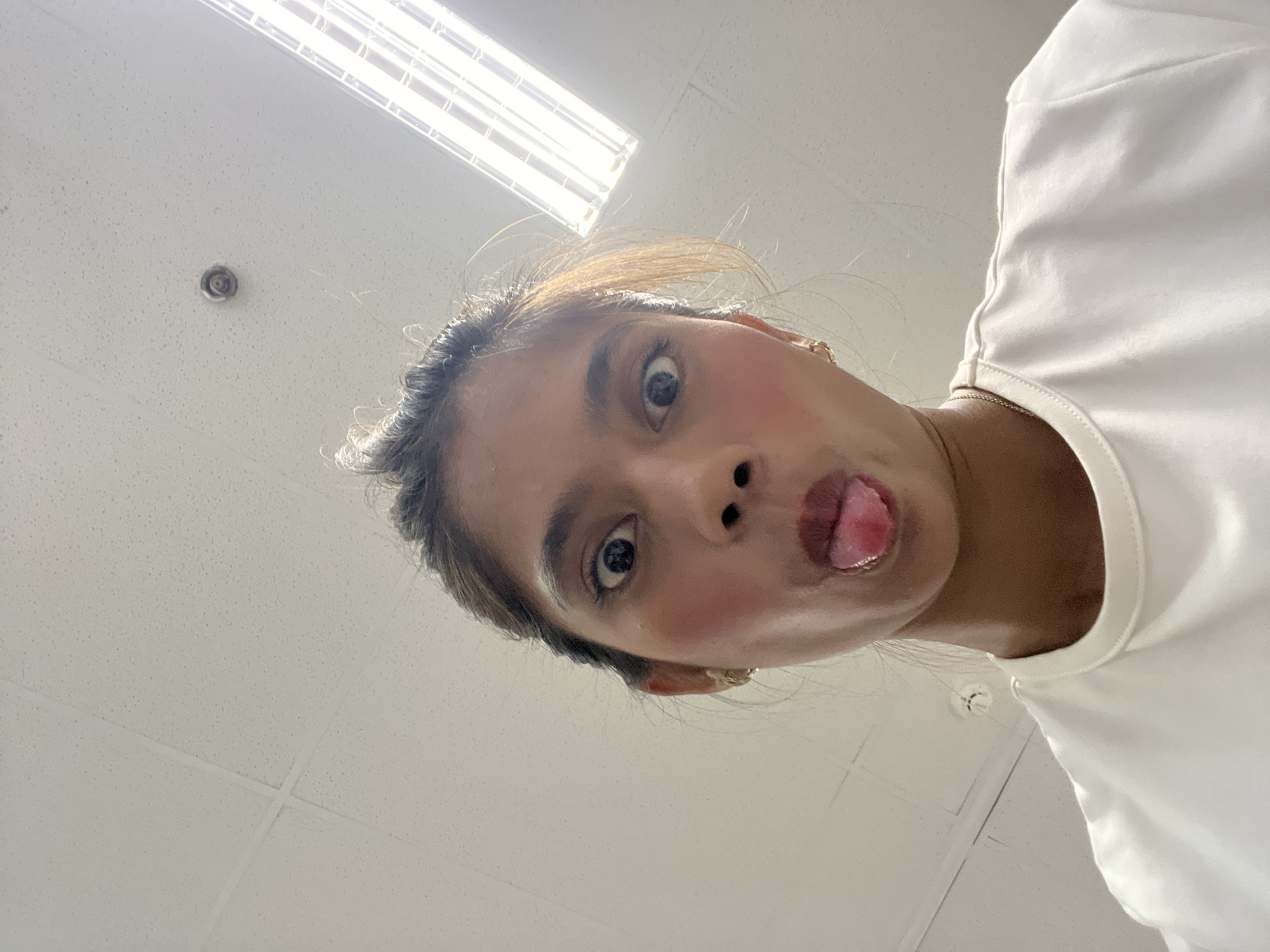 A young woman is seen in an upward-facing, close-up perspective. Her face is central, with her tongue sticking out slightly. She has light brown skin, dark eyes, and dark hair pulled back. She is wearing a white shirt. The background is an interior space with a white ceiling panel and a fluorescent light fixture visible in the upper left. A circular ceiling fixture is also visible. It is not possible to determine if the image was taken in Davao City.