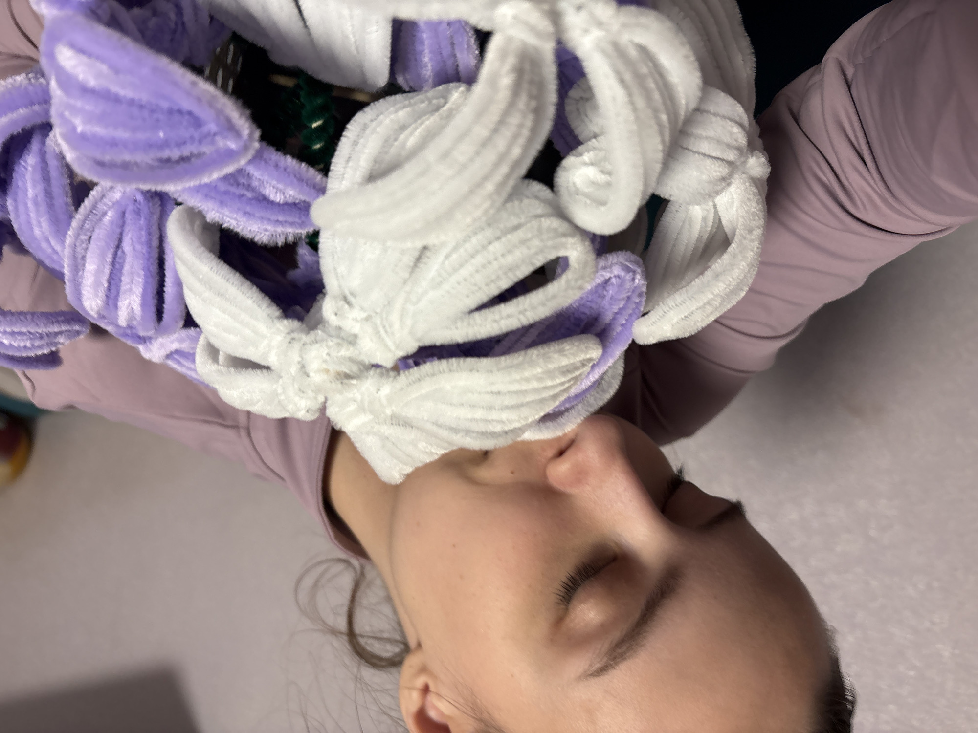 A close-up, tilted image shows a young person interacting with a bouquet of artificial flowers made from pipe cleaners. The person, visible from the nose downwards, has fair skin and closed eyes, suggesting a moment of calm or contemplation. Dark hair is visible near the bottom of the frame. They are wearing a light purple long-sleeved top.
The bouquet, which occupies a significant portion of the upper and central frame, is composed of numerous flowers crafted from fuzzy pipe cleaners. These flowers are primarily white and light purple, featuring a textured, soft appearance. Small dark green elements, possibly stems or leaves, are faintly visible within the arrangement. The person's nose is in close proximity to the white and purple flowers, creating an impression of smelling or appreciating the craft.
The background is a soft, light-colored surface, likely an indoor floor or table, with some indistinct darker areas and shadows. There is no visible text in the image. The scene conveys a peaceful interaction with a handmade item. The location context is Прикамский, Russia.