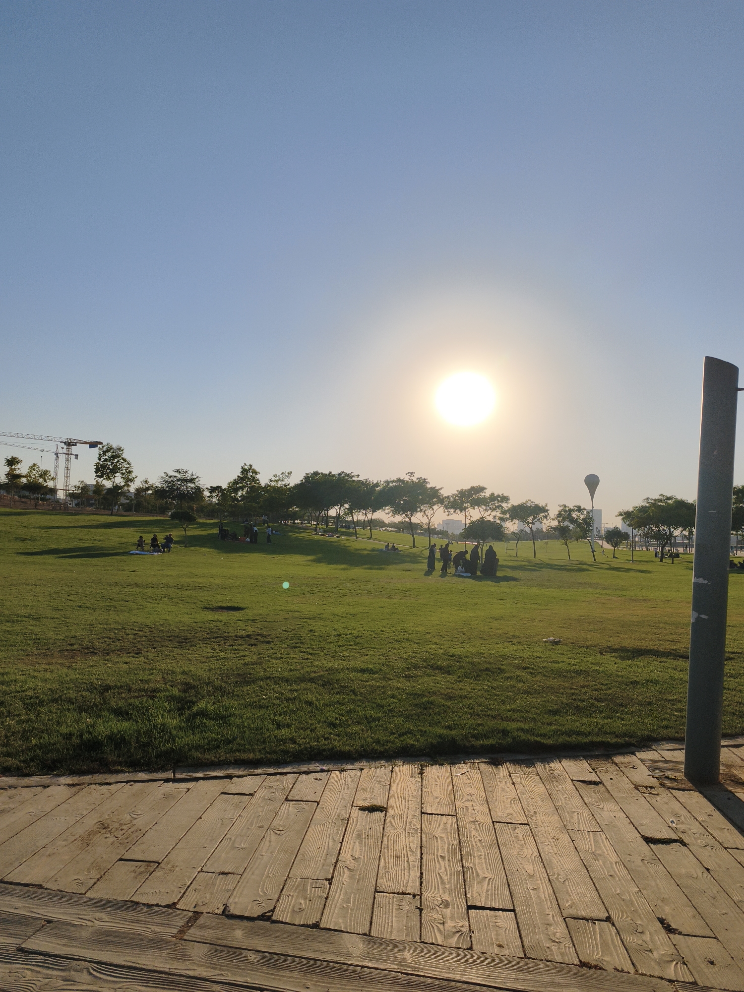 The image depicts a spacious, well-maintained park in Be'er Sheva, Israel, under a clear, bright sky with the sun prominently positioned. The foreground features a wooden pathway with a light brown, weathered appearance, laid out in planks. Beyond the pathway lies a vast expanse of vibrant green grass, sloped gently upwards towards the background.
Scattered across the lawn are several groups of people, suggesting a leisurely afternoon. Some individuals are seen sitting or lying on blankets, possibly picnicking or relaxing. One group in the mid-ground appears to be engaged in conversation or an activity together. The trees in the park are a mix of leafy and sparse varieties, casting distinct shadows on the grass due to the bright sunlight.
In the background, construction cranes are visible on the left side of the frame, indicating ongoing development or infrastructure projects in the vicinity. Further in the distance, a distinctive balloon-like structure atop a tall, white building stands out against the horizon. The overall atmosphere is one of tranquility and outdoor recreation. The lighting suggests it is either late afternoon or early morning.