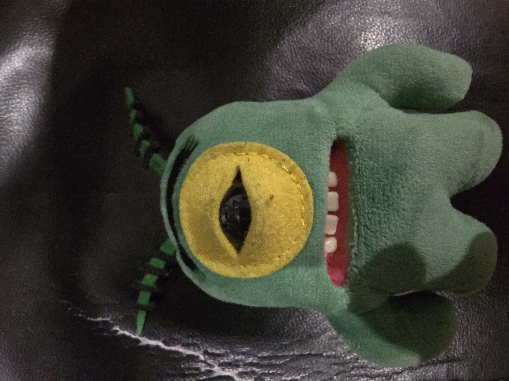A green plush toy, resembling the character Plankton from the Spongebob Squarepants cartoon, is positioned vertically against a dark, textured surface. The toy has a large, circular yellow eye with a black pupil that is slightly off-center. Its mouth is open, revealing white teeth and a pink interior. Two antennae, adorned with green and black stripes, protrude from the top of its head. The background is a dark, possibly leather, surface with subtle wrinkles and lighter streaks, suggesting it might be a couch or a similar piece of furniture. The lighting creates soft shadows around the toy. The location is unknown.