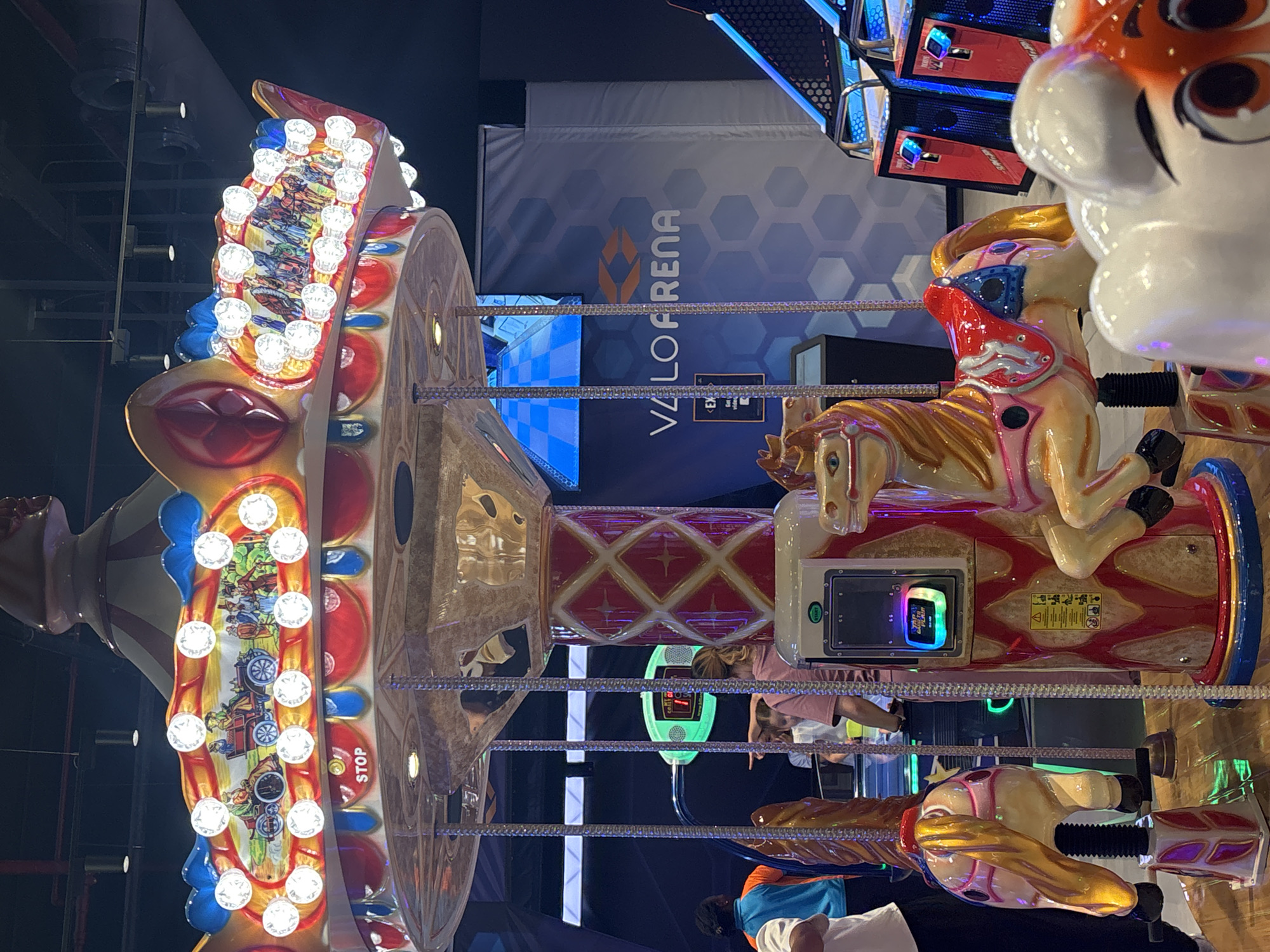 The image captures a carousel inside a Valo Arena entertainment venue, likely located in Dubai, United Arab Emirates. The carousel features brightly colored horses with detailed saddles and ornate poles. Above the carousel, a decorative canopy is adorned with lights and painted scenes depicting vintage vehicles and figures. A "STOP" sign is visible on the edge of the canopy. In the background, arcade games with illuminated screens are present, along with a large banner displaying the "VALO ARENA" logo. Several people, including children and adults, are visible around the carousel and in the surrounding area, some appearing to be playing or observing the rides. The overall environment suggests a lively indoor amusement or arcade setting.