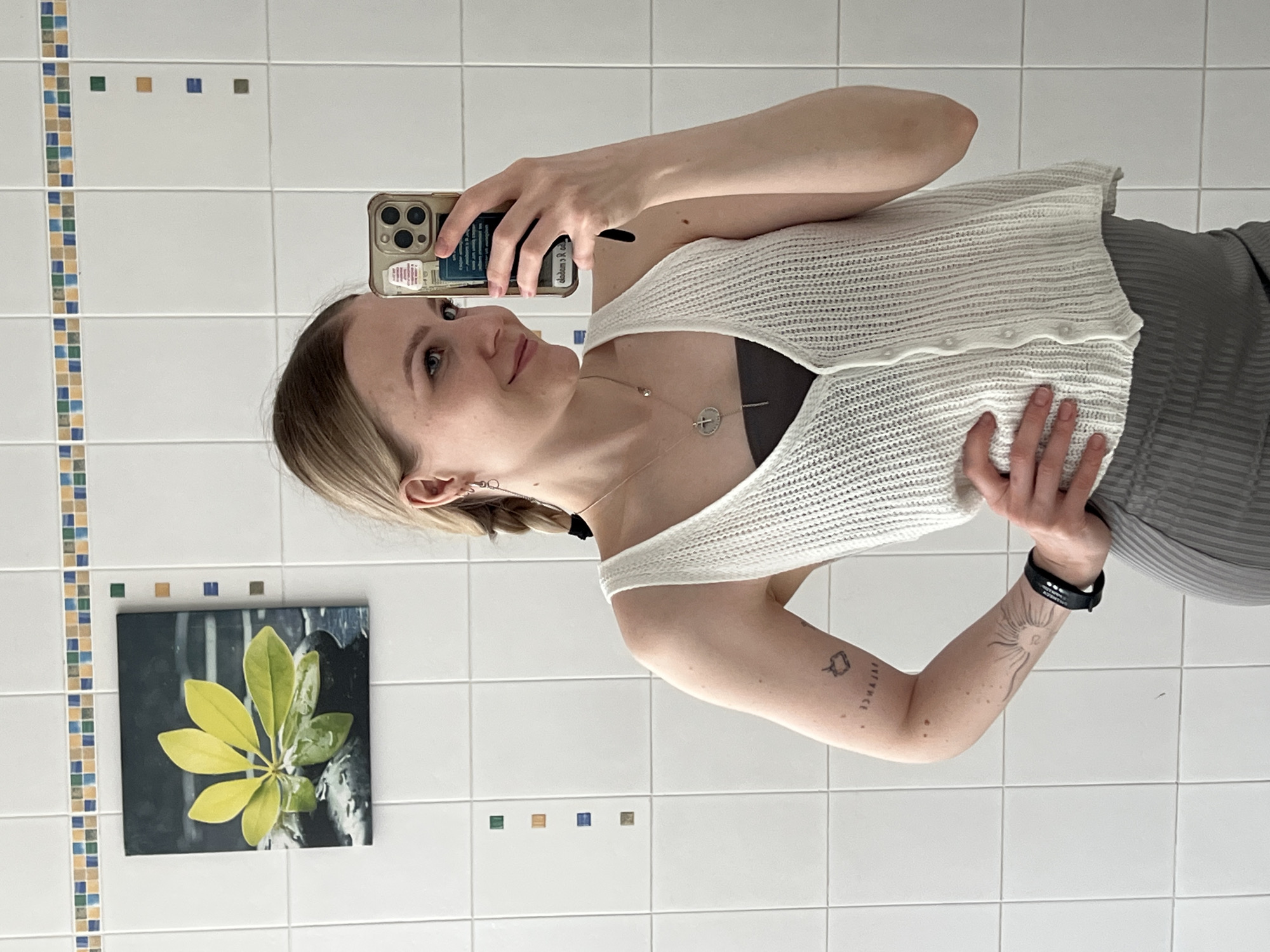 A fair-skinned female with blonde hair is taking a selfie in a bathroom with white tiled walls. The bathroom has a decorative row of colorful tiles at the top and a small picture of a green leaf against a dark background. The woman is wearing a white, knitted vest over a dark top and a grey skirt or pants. She is holding a smartphone in her left hand and is looking at the screen. She has visible tattoos on her left forearm and is wearing a black wristband. The photograph does not contain information to determine a location in Khimki, Russia.