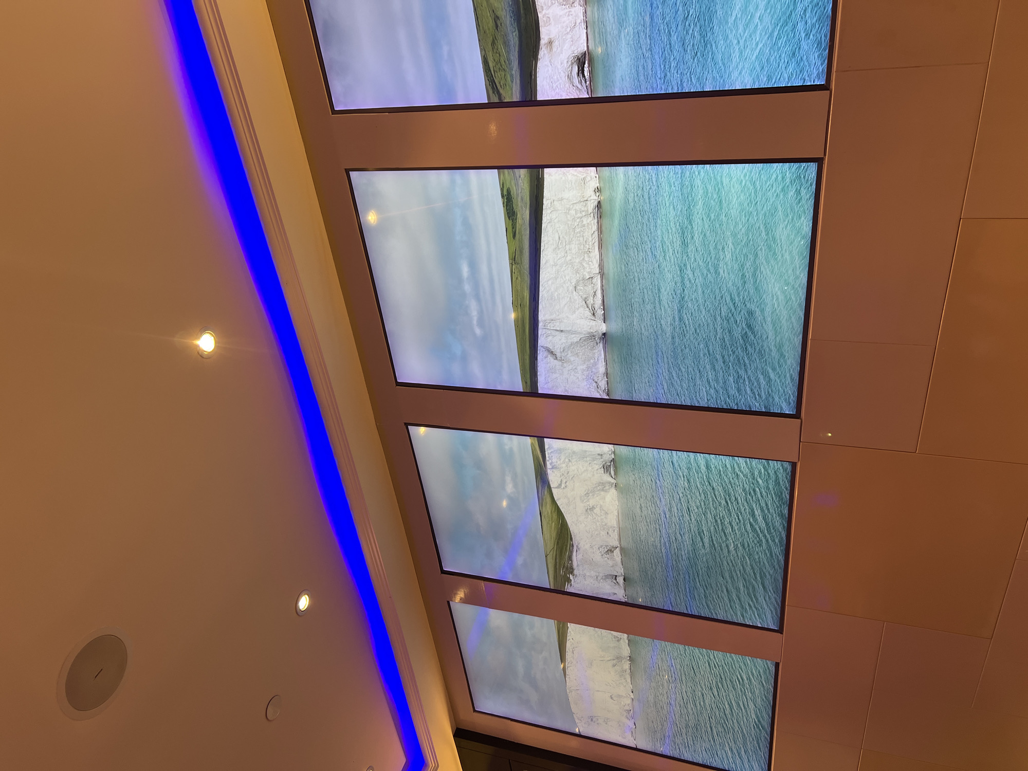 Four large, vertical digital screens display a scenic view of white cliffs meeting a turquoise ocean under a cloudy sky. The screens are mounted side-by-side on a warm, beige wall. A strip of blue LED lighting runs along the top edge of the wall, casting a glow onto the ceiling. The ceiling is cream-colored and features recessed lighting and a round speaker. The setting appears to be an indoor space, possibly a hotel lobby or a retail environment, given the lighting and display setup. No people are visible in the image. This installation is located in Sharm El Sheikh, Egypt, likely within a commercial or entertainment venue.