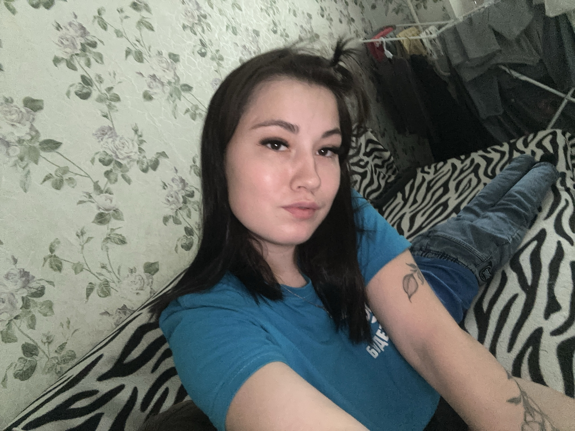A young woman with long, dark hair is lying on a bed with a zebra-print comforter. She is wearing a blue t-shirt and blue jeans. She has a tattoo of a fruit on her left arm and another tattoo on her right arm that is partially visible. The background appears to be a wall with a floral pattern. The scene is indoors, likely in a residential setting. The image is a selfie, taken from a slightly high angle. The location of Chaikovsky, Russia, is not depicted in the image.