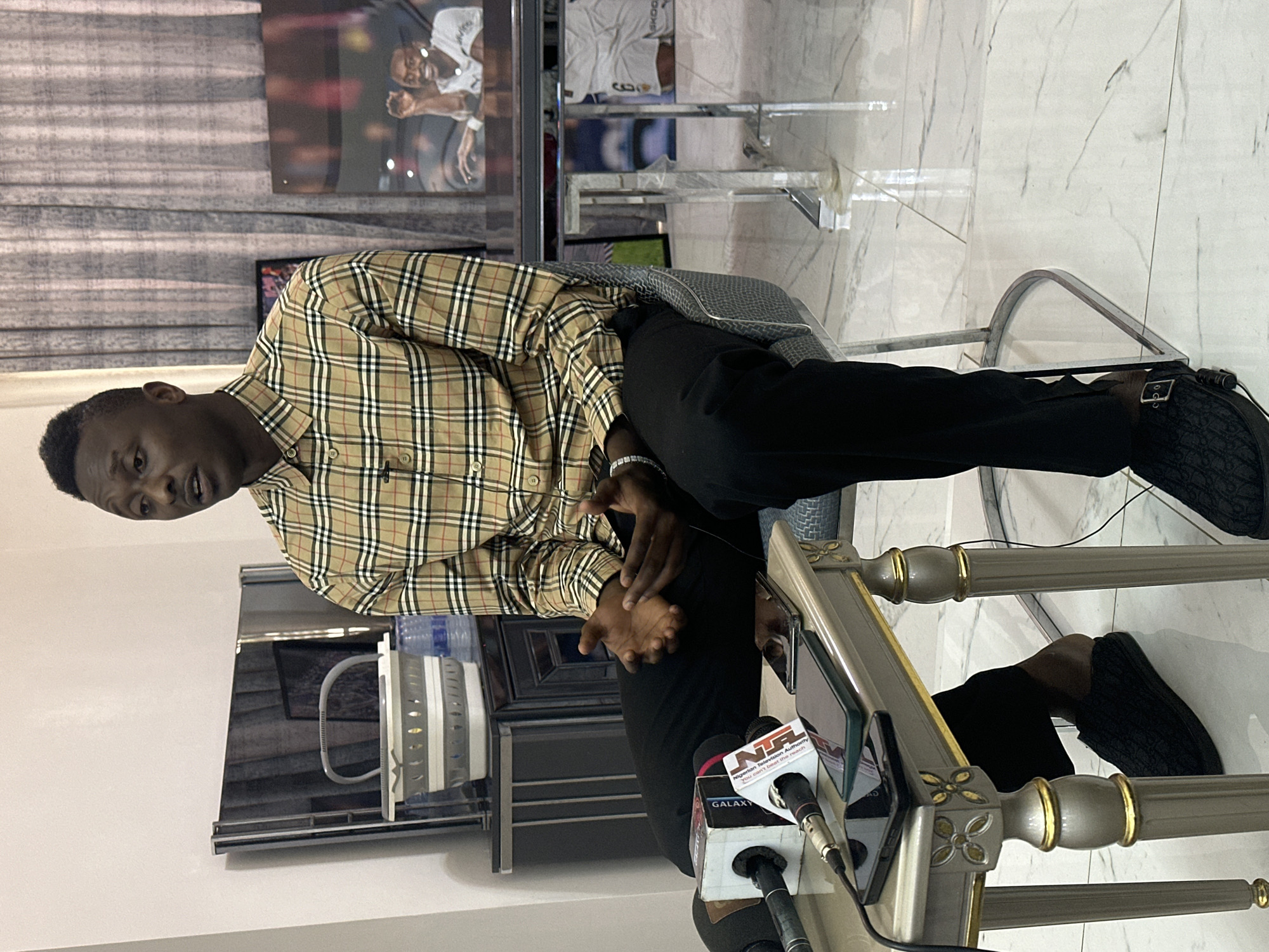 A man, likely of African descent, is seated and being interviewed in what appears to be a well-lit indoor setting. He is wearing a tan, plaid button-down shirt with a black and white checkered pattern and black trousers. His hands are clasped in front of him as he looks towards his left, his expression neutral. A microphone with the "NTA Nigerian Television Authority" logo and the tagline "You can't beat the reason" is positioned in front of him, along with other microphones and what appears to be a tablet device.
The background reveals a modern interior with a marble-tiled floor and walls. A television screen is visible, displaying an image of two individuals with their arms raised in celebration. Behind the man, there's a curtain and a glimpse of more television screens and shelving units. A water dispenser with several bottles is also present. The scene suggests a press conference or interview event. The lighting indicates it is daytime. The location context provided is Unguwar Sarki, Nigeria.