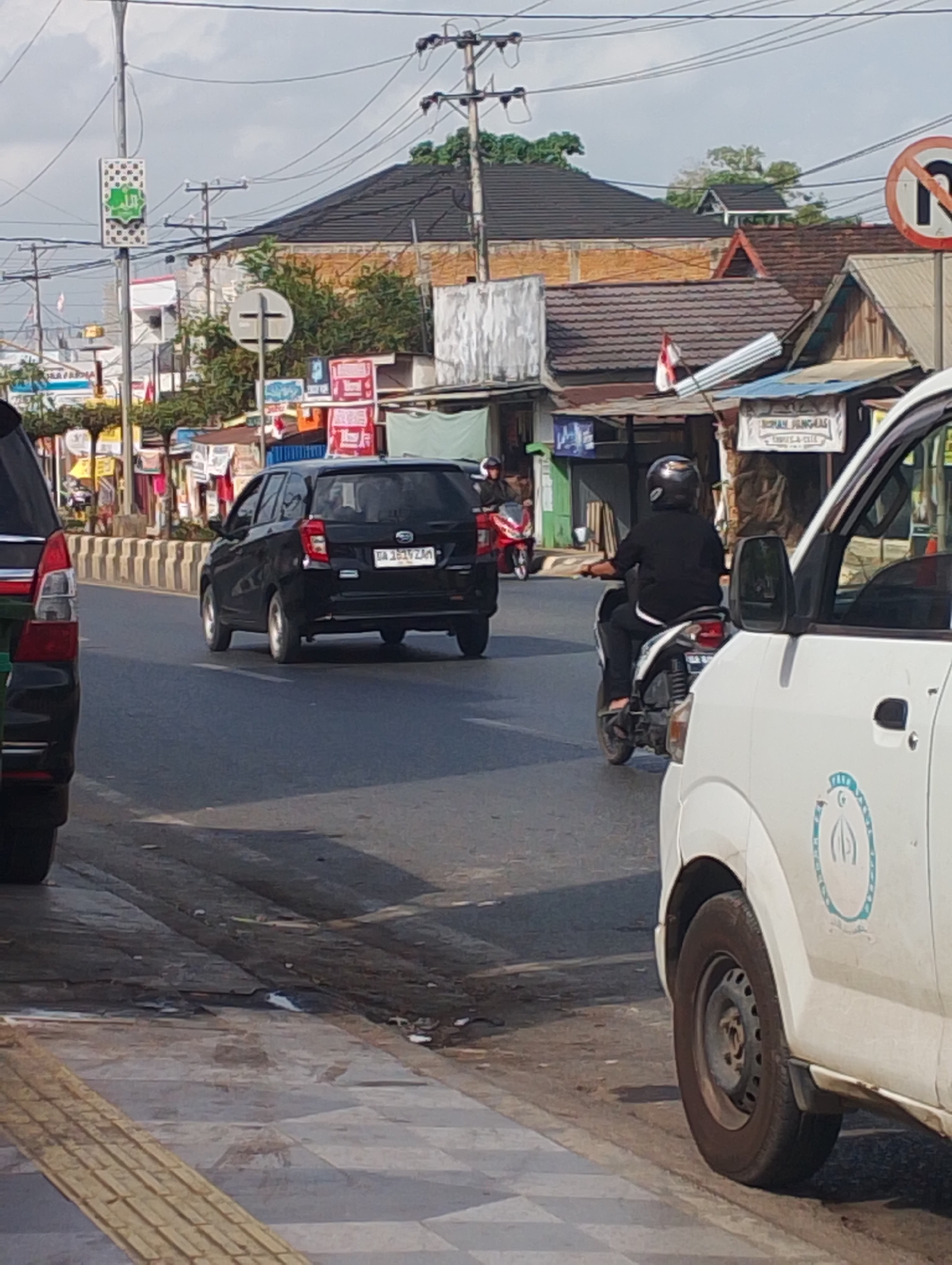 The image depicts a street scene in Baroqah, Indonesia. A black car with the license plate "GA 1819 ZAH" is driving on the road. In front of it, a person on a motorcycle wearing a black helmet and black clothing is also moving. The road is paved, and there is a sidewalk with yellow tactile paving. Buildings line the street, some with tiled roofs and awnings. Overhead, power lines crisscross the sky, and there are several street signs, including a circular sign with a red border and a diagonal line through a 'no U-turn' symbol. A white vehicle is partially visible in the foreground on the right side of the image. The sky is partly cloudy.