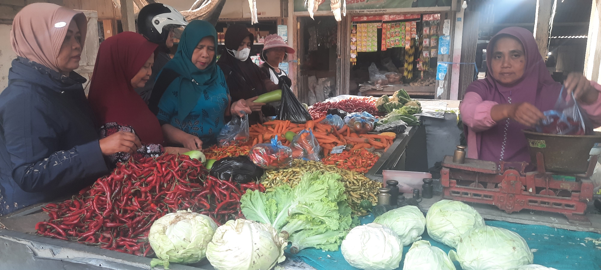 This image depicts a vibrant street market scene in Selopuro, Indonesia, featuring several women vendors actively engaged in selling produce. The foreground is dominated by a large display of red chilies, with cabbages and lettuce also visible. A variety of other vegetables, including carrots and smaller peppers, are arranged on a table. In the background, a storefront displays a selection of packaged goods, suggesting a bustling commercial area. One woman, wearing a purple hijab and pink top, is in the process of weighing produce on a vintage red scale, indicating a transaction is underway. Another woman in a teal hijab is seen handling a long green vegetable. The overall atmosphere is one of an active marketplace, with the vendors showcasing their fresh offerings to potential customers.