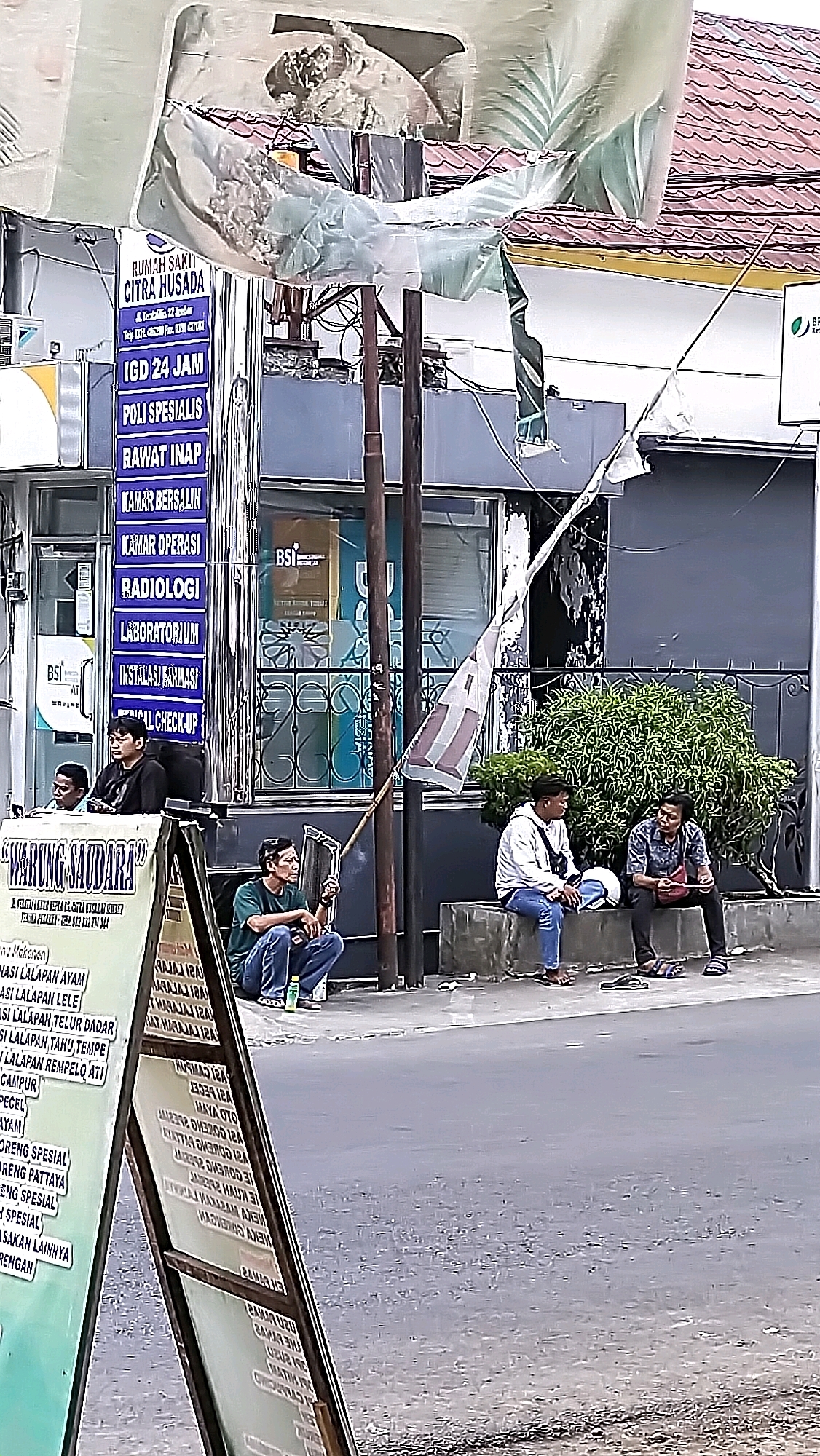 The image depicts a street scene in Jember, Indonesia, likely in front of a medical facility and a food stall. In the foreground, a triangular sign displays text in Indonesian, featuring the name "WARUNG SAUDARA" and a menu. Behind this sign, several individuals are present. A man in a green shirt is seated on the ground, holding a stick. Two other men are seated on a low wall to the right; one wears a white hooded jacket and blue jeans, while the other is in a patterned shirt and dark pants. Further back, a blue sign with white text lists various medical services offered by "RUMAH SAKIT CITRA HUSADA," including "IGD 24 JAM," "POLI SPESIALIS," and "KAMAR INAP." The building facade behind the sign has windows with an BSI logo visible. The environment includes a paved street, a sidewalk, a building with a tiled roof, and dense green foliage. A tattered flag is also visible, attached to a pole. The overall impression is a snapshot of daily life in a commercial area.