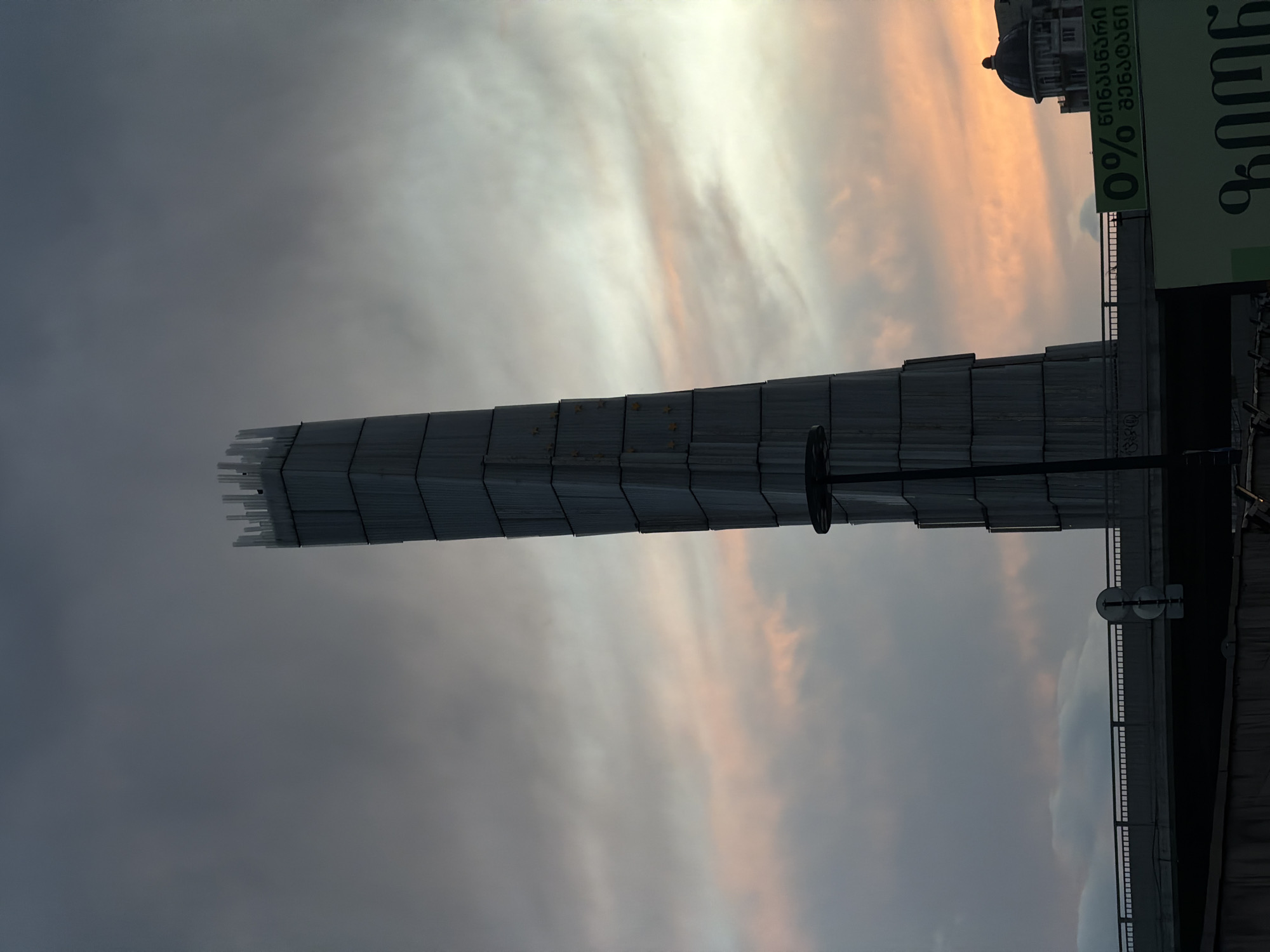 This image shows an upward view of a tall, rectangular structure composed of multiple segments or blocks, likely part of a building or monument, under a cloudy sky with hints of sunset hues in the city of Tbilisi, Georgia. The sky is a mix of grey and light orange shades. To the right, parts of a building with a dome and a green advertisement sign with Georgian writing are visible. Also present are a traffic sign and a railing that seem to be part of a bridge or elevated walkway. The photo appears to be taken from below, capturing the structure against the sky.
