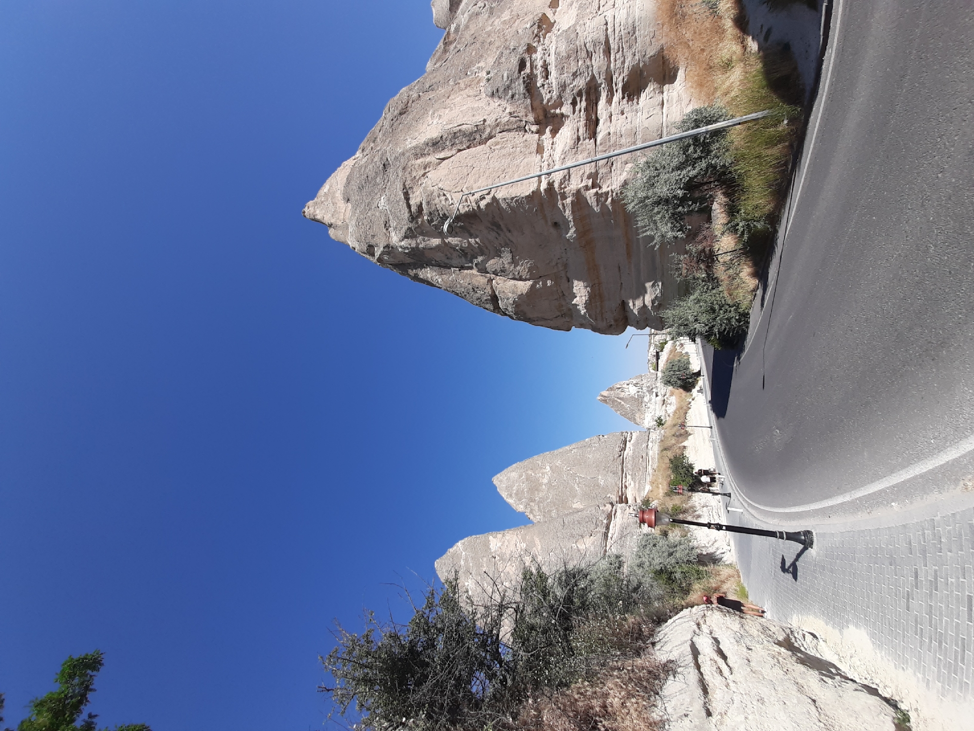 The image depicts a scenic road with unique rock formations in Göreme, Turkey, a region known for its fairy chimneys. A paved sidewalk runs alongside the road on the left, bordered by a rough, light-colored rock face. A lone woman wearing a dark dress and a red hat leans against the rock face on the sidewalk, facing away from the camera. A vintage-style lamppost stands on the sidewalk near the woman. Several other people, appearing as small figures, walk further down the sidewalk. The road itself is asphalt and curves gently to the right, leading into the distance. In the background, the distinctive cone-shaped rock formations, characteristic of Göreme's landscape, rise against a clear, vibrant blue sky. Sparse, dry vegetation and some trees dot the areas around the rock formations. The overall scene is one of natural beauty and a popular tourist destination.