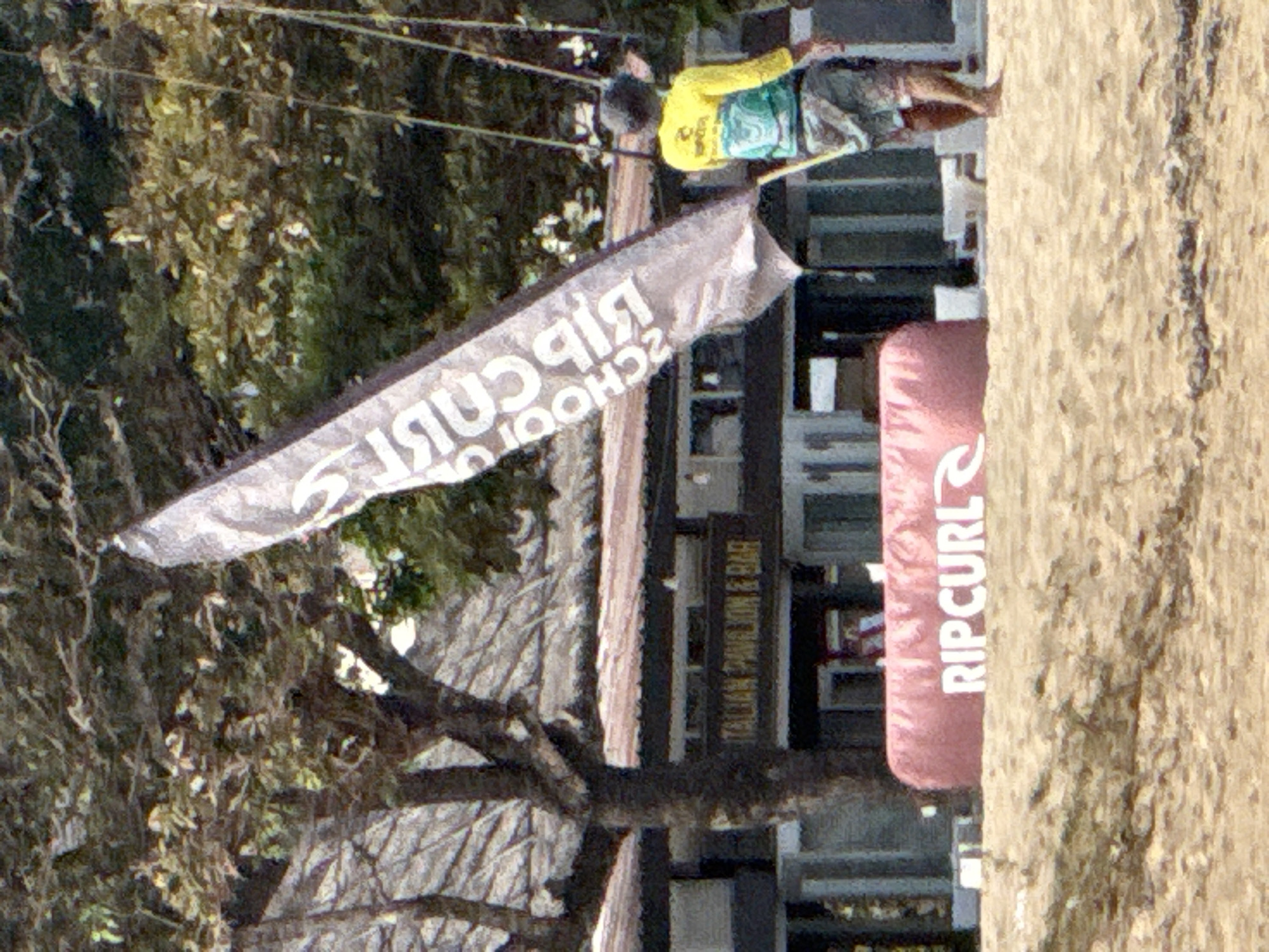 The image captures a scene on a sandy beach in Denpasar, Indonesia, featuring a building with signage that reads "ITALIAN PAVILION & BAR." In the foreground, a large pink inflatable object with the "RIPCURL" logo is visible. A flag displaying "RIPCURL SCHOOL" is prominently featured, caught by the wind. To the right, a person is seen from the back, wearing a yellow and blue shirt and patterned shorts, holding a pole. The background consists of trees and foliage, with the beach sand dominating the lower portion of the image.