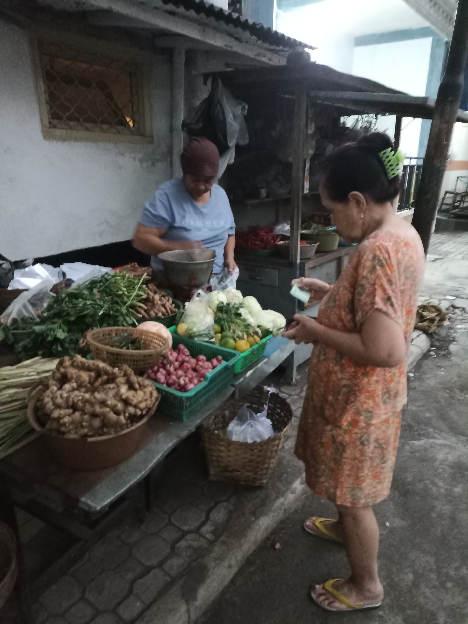 The image shows two women at an outdoor market stall in Lumajang, Indonesia. The woman on the left, wearing a blue t-shirt and headscarf, is behind the stall and appears to be weighing or preparing produce. The stall is laden with various fresh vegetables, including leafy greens, ginger, shallots, and what appear to be lemons and cabbage. The woman on the right, dressed in an orange floral print dress and yellow flip-flops, is facing away from the camera and examining her mobile phone. The market stall has a corrugated metal roof supported by wooden posts, and the ground is paved with cobblestones. The overall lighting suggests it is either early morning or late afternoon.