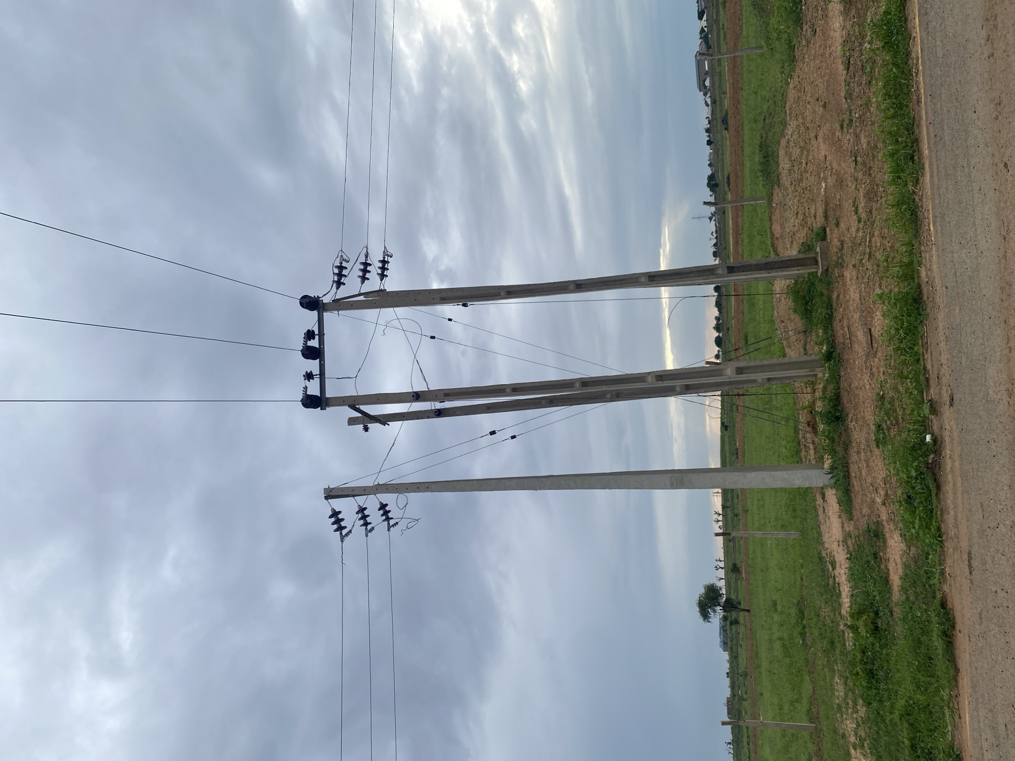 The image shows a rural landscape with power lines stretching across the frame. Three concrete utility poles are visible, supporting the electrical wires. The sky is overcast with dramatic clouds, suggesting it might be a cloudy day or that a storm is approaching. The ground is a mix of green grass and dry, patchy soil, indicating a natural environment. In the background, there are some trees and a few buildings, hinting at a sparsely populated area. The perspective is from ground level, looking upwards at the power lines and sky, with a road or dirt path visible on the right side of the frame. There are no people or specific activities depicted, making it a static scene of infrastructure in a natural setting. The overall mood is somewhat somber due to the heavy clouds, but the greenery suggests life and growth. The location context of Dumnga, Nigeria, aligns with the visual cues of a rural African landscape.