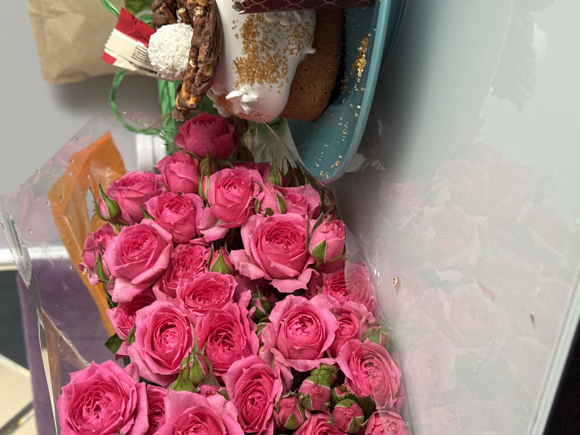 A still life features a bouquet of pink roses in the foreground, wrapped in clear plastic, with a gift basket containing what appears to be a pastry, covered in white frosting and gold sprinkles, placed above the flowers. The background includes a light-colored wall and a glimpse of a paper bag. The setting and location are unidentified.