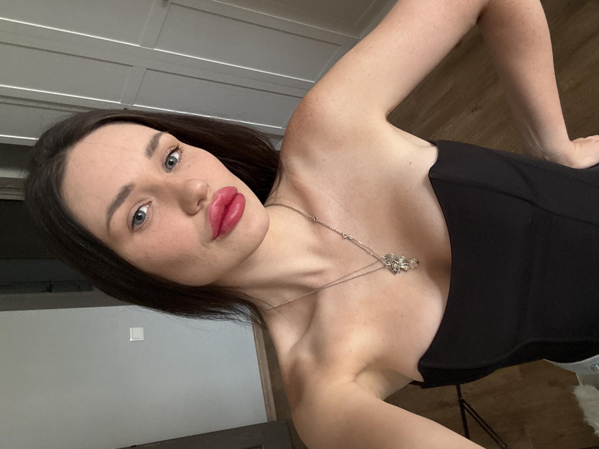 A young woman with dark hair and blue eyes is taking a selfie in what appears to be an indoor setting. She is wearing a strapless black top and a silver necklace with a charm. Her lips are full and colored bright pink. She has freckles visible on her shoulders and arms. The background consists of a light gray wall with a white electrical outlet, and a closet with white paneled doors. The flooring is wooden. The lighting suggests it is daytime. The image does not provide any specific indicators of the location being Tbilisi, Georgia.