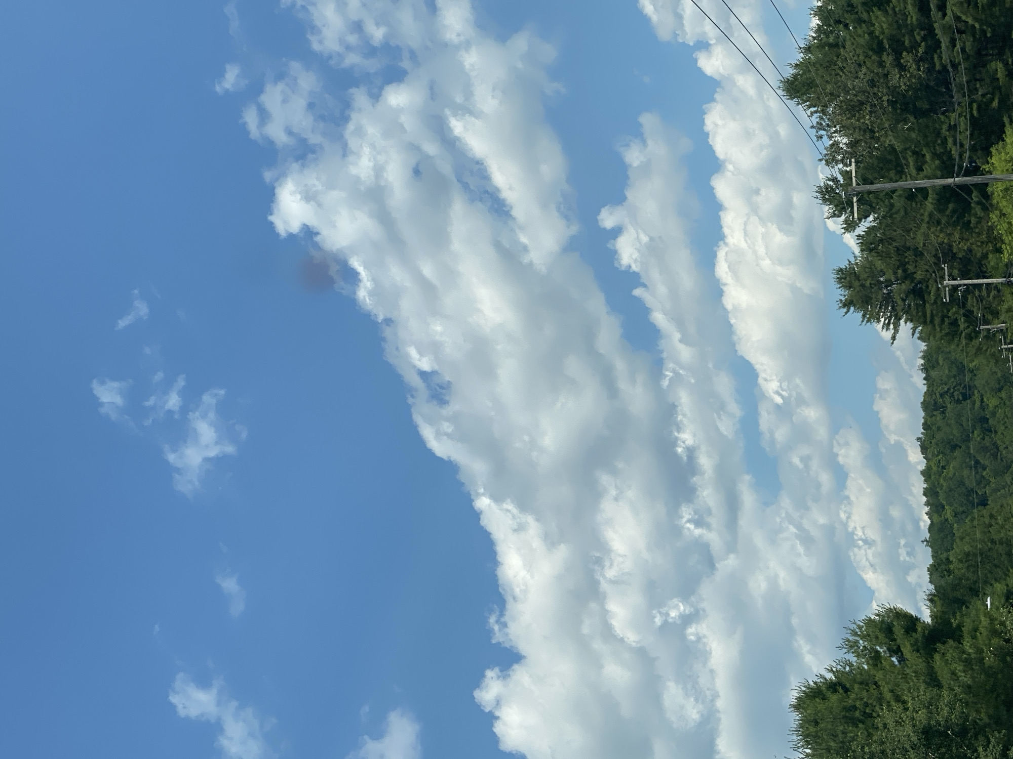 The image shows a bright blue sky filled with fluffy white clouds. A small, dark, reddish-brown smudge is visible in the upper right quadrant of the sky, possibly smoke. The lower portion of the image features a dense line of evergreen trees, and in the foreground, two wooden utility poles with wires strung between them are visible, with the tops of other poles and wires peeking through the trees. The scene appears to be taken from a roadway, as indicated by the telephone poles and wires. The image was captured in Lac-Simon, Canada.