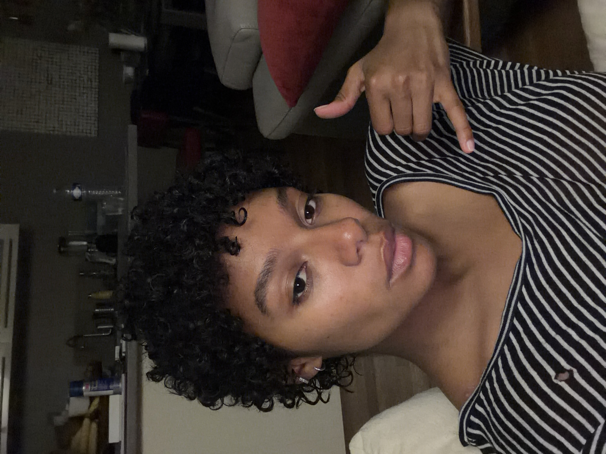 A person with curly, dark hair is looking at the camera. They are wearing a black and white striped shirt and pointing at themselves with their right hand. The background is dimly lit, showing a gray chair with a red pillow and a shelf with various items on it. There is no indication of the city of Champs-sur-Marne, France, or any evidence to suggest this image was taken there.
