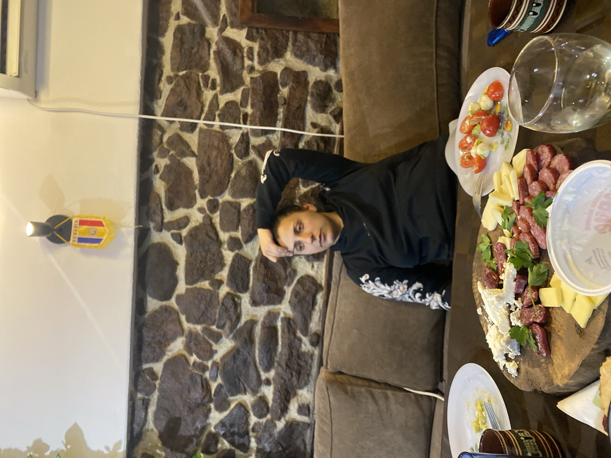 A young person reclines on a brown sofa in what appears to be a home or restaurant setting. They are positioned next to a laden table, which has a wooden platter containing various types of cheese, salami, and greenery. The table also holds a salad, a glass, and various dishes. In the background, a wall is adorned with stone cladding. There is also an elongated Romanian flag in the background. The precise location is Belopoptsi, Bulgaria.