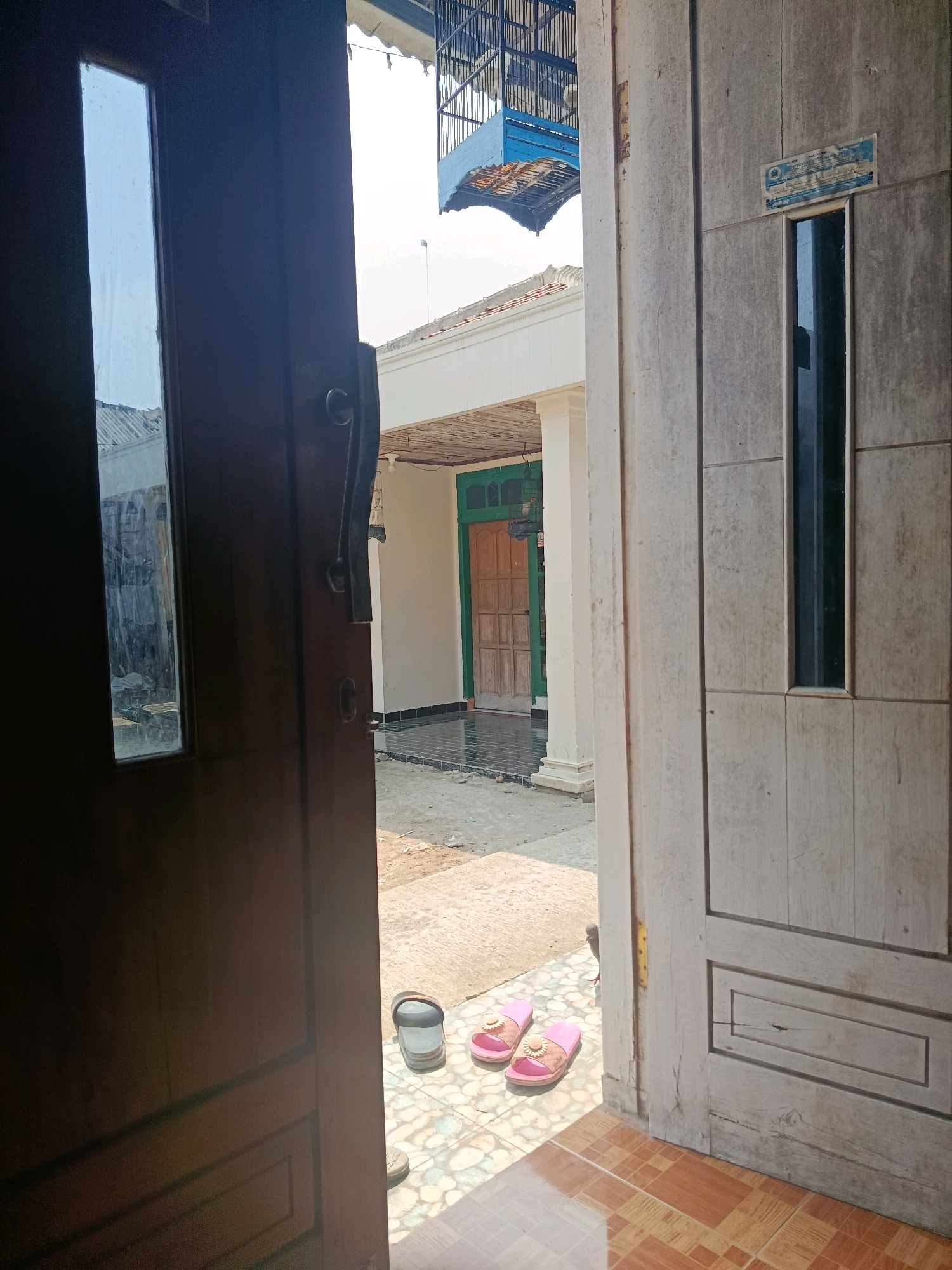 The image depicts an open doorway leading to a courtyard in Binangun, Indonesia. On the left, a dark wooden door is ajar, revealing a partially visible reflection in its glass pane. To the right, a light-colored wooden door stands open, showcasing its textured surface. In the foreground, a pair of pink slippers lies on a tiled floor. Beyond the doorway, the courtyard features a paved area with scattered debris, a bucket, and more colorful tiles. In the background, a building with a white facade and a green-framed wooden door is visible. Overhead, a blue birdcage hangs from a light-colored awning. The scene is illuminated by bright daylight, suggesting an outdoor setting.