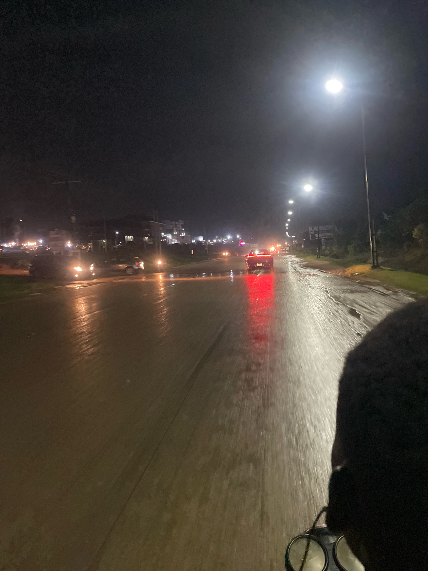The image depicts a nighttime scene in Galadimawa Village, Nigeria. The road is wet and reflects the lights from street lamps and vehicles. Several cars are visible, with one car in the foreground having its red taillights illuminated, casting a strong reflection on the wet road. The perspective suggests the photo was taken from a moving vehicle, possibly a motorcycle, as a dark, silhouetted shoulder and head are visible on the right side of the frame. In the background, buildings and more streetlights indicate an urban or semi-urban environment. The overall atmosphere is one of a rainy night on a road in Nigeria.