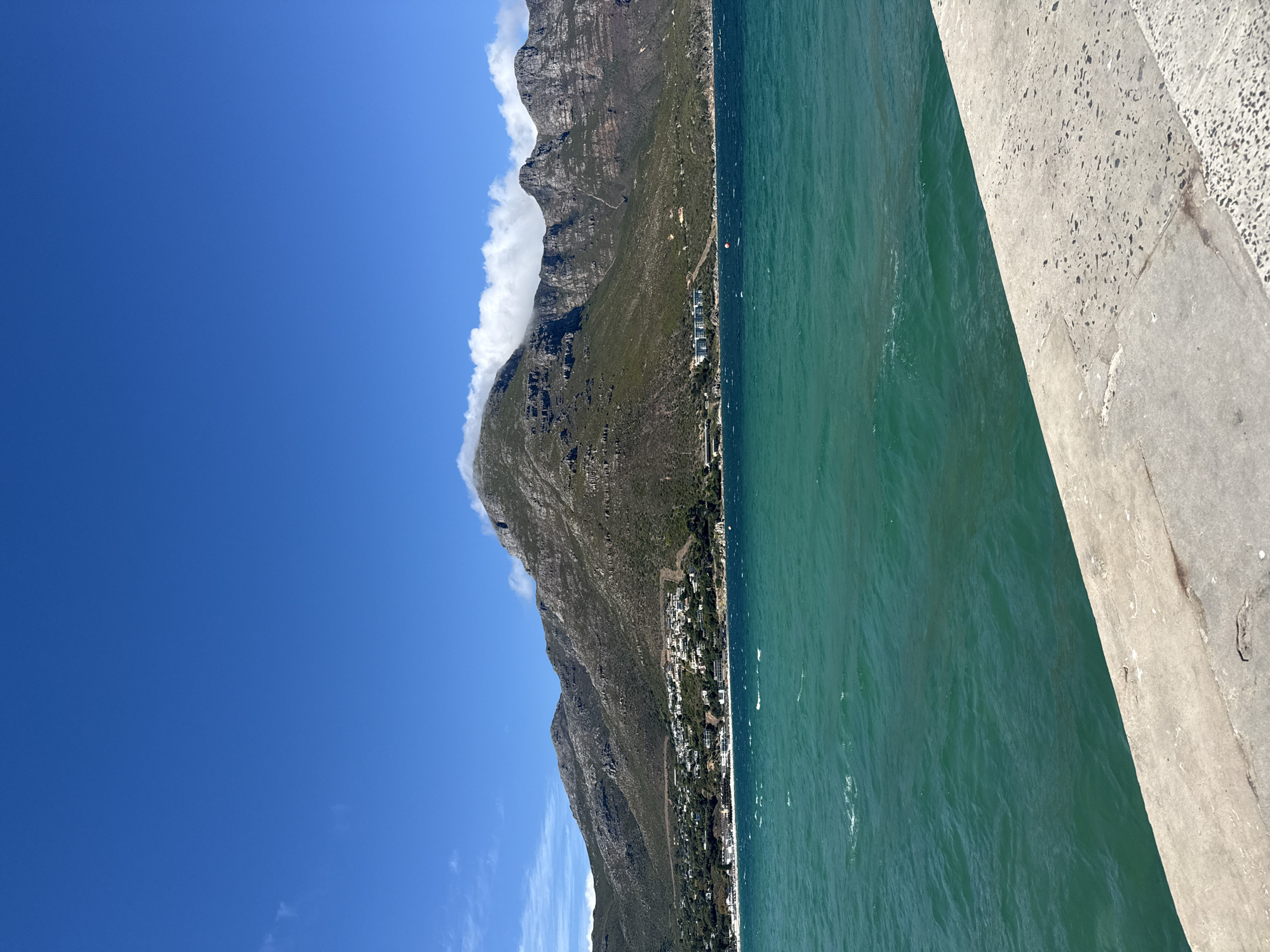 The image depicts a coastal view in Hout Bay, South Africa, featuring a mountain range partially obscured by clouds on the left side of the frame, a strip of coastline with buildings running along the mountain’s base, and the turquoise ocean to the right. A concrete structure is visible in the foreground, possibly part of a pier or seawall. The sky is clear blue. No people are visible.