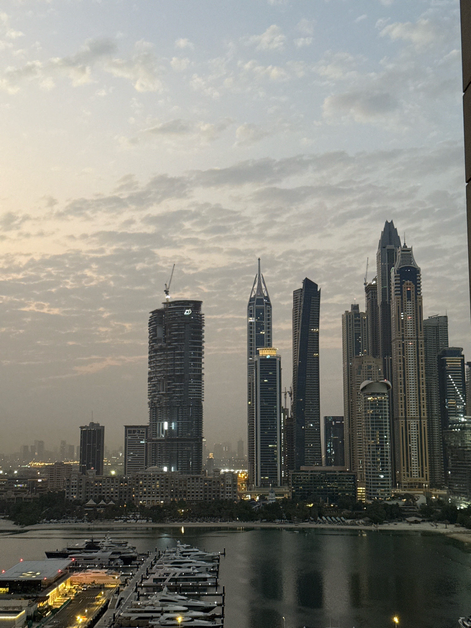 The image shows a panoramic view of the Dubai Marina in Dubai, United Arab Emirates. The foreground features a marina filled with numerous luxury yachts docked at piers. A marina building with illuminated lights is also visible on the left. In the midground, a waterfront promenade lined with trees and buildings stretches along a sandy beach. The background is dominated by a dense cluster of modern skyscrapers, characteristic of Dubai's iconic skyline. The sky above is overcast with scattered clouds, suggesting either early morning or late evening. The overall scene depicts a vibrant and upscale urban waterfront environment.