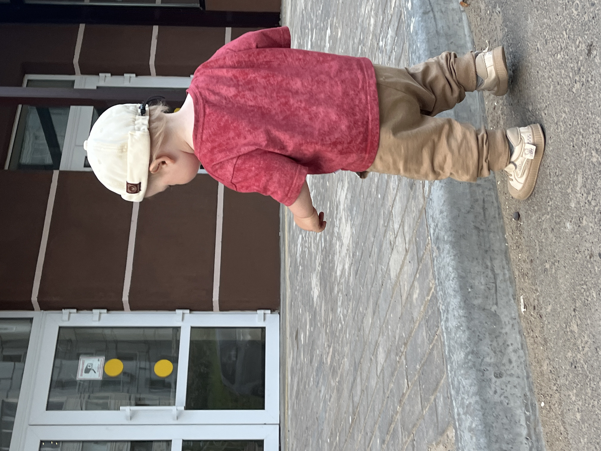 A young child wearing a white cap, red t-shirt, tan pants, and tan sneakers is walking on a brick sidewalk adjacent to a building. The building has white framed windows and doors. The windows have yellow circles on them. The location is in the city of Chaikovsky, Russia.