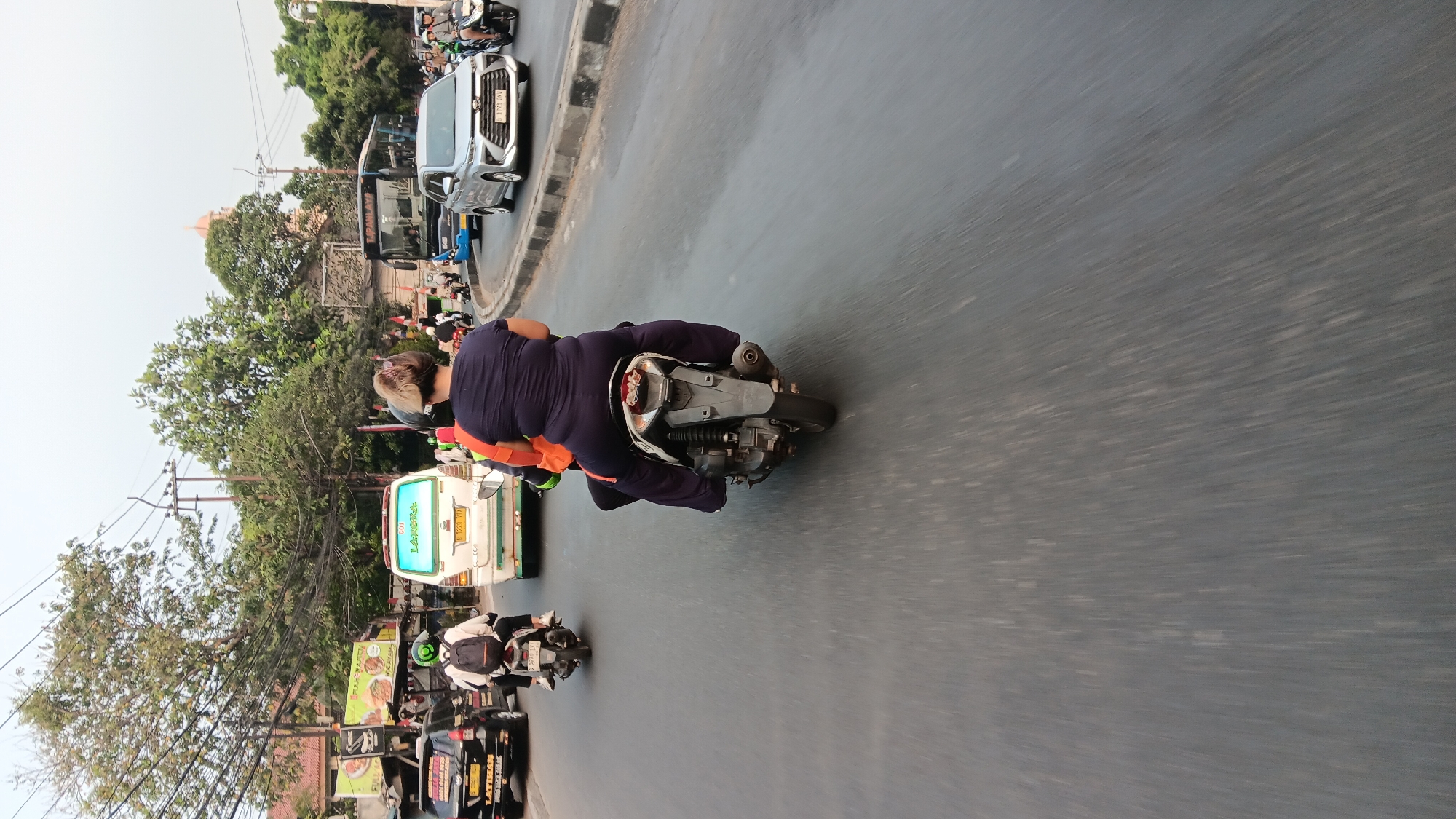 A first-person perspective shot captures a busy street scene in Tangerang, Indonesia, where traffic is flowing. The foreground features a motorcycle from behind, with a passenger wearing a dark blue shirt and dark pants. The passenger's hair is pulled back, and they are seated behind the driver, who is also wearing a helmet and dark clothing. The motorcycle itself is a dark color, with visible rear suspension and an exhaust pipe.
In the middle ground, other vehicles are visible on the asphalt road. To the left, a black car is seen from the side, with a license plate that reads "B 1239 VTA". A person on a motorcycle, wearing a green helmet and a backpack, is to the left of this car. Further ahead, a white van is driving, displaying "601 LARONA" on its rear. To the right of the van, a silver car with a license plate "B 1791 JAU" is in motion. Several other motorcycles and a blue bus are also present in the traffic.
The background reveals a street lined with trees and buildings. Electrical wires are strung across utility poles. The sky is overcast, suggesting either an early morning or late afternoon. The overall impression is one of a typical urban environment with active traffic and pedestrian activity, consistent with a location like Tangerang, Indonesia.