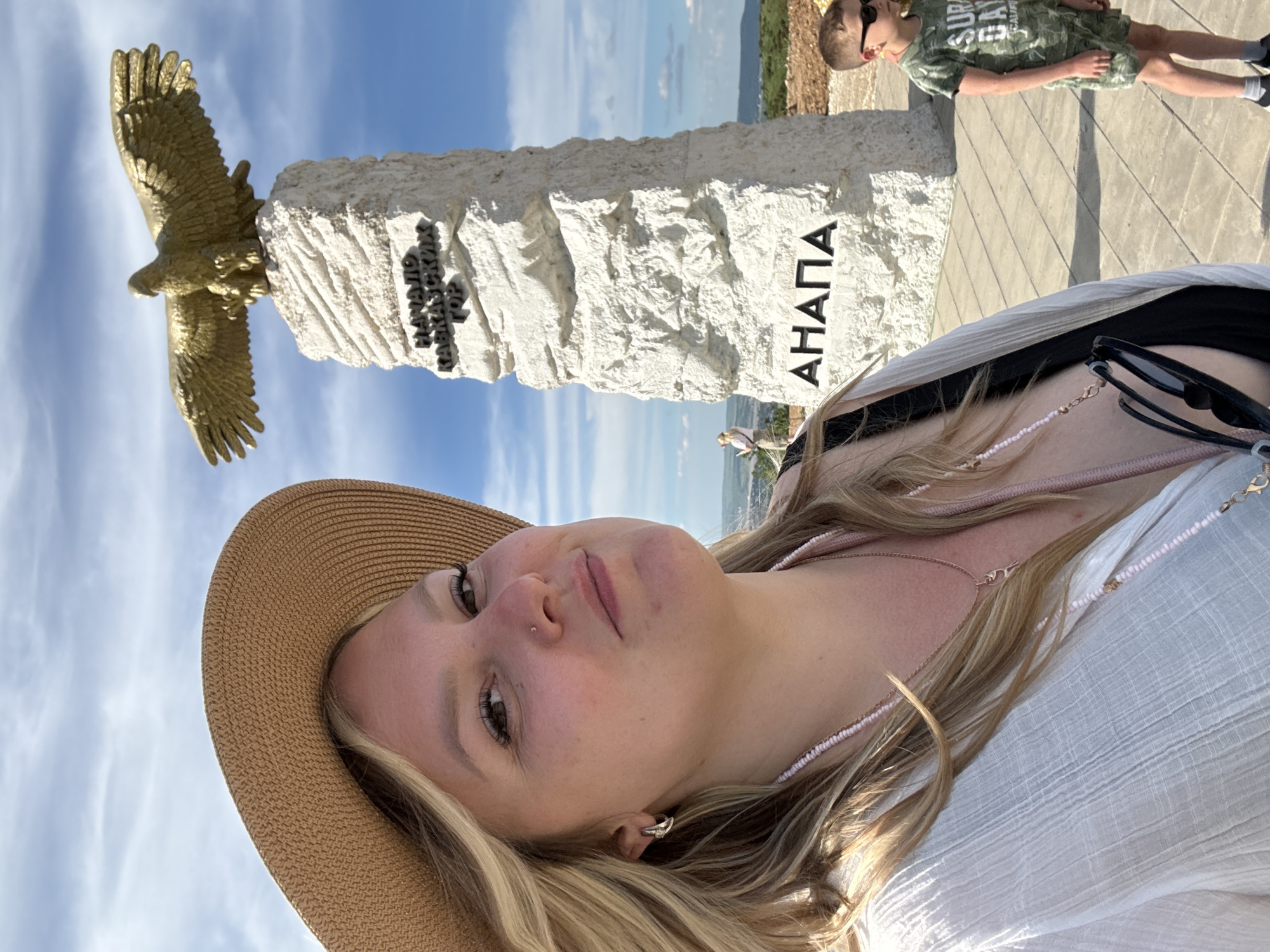 The image captures a selfie-style portrait of a woman in the foreground, with a monument and a child visible in the background. The woman, with blonde hair, wears a wide-brimmed straw hat and a white, textured top, along with a delicate beaded necklace and sunglasses hanging from her neck. She looks towards the camera with a subtle expression.
Behind her stands a significant monument composed of a large, rough-cut white stone stele topped with a golden statue of an eagle in flight. The monument features Cyrillic inscriptions. On the left side, the text "НАЧАЛО КАВКАЗСКИХ ГОР" (START OF THE CAUCASUS MOUNTAINS) is visible. On the right side of the stele, the word "АНАПА" (ANAPA) is clearly inscribed in large black letters.
To the right of the monument, a young boy in a green camouflage-patterned t-shirt, shorts, and sunglasses stands on a paved area, seemingly looking out at the distant landscape. The setting appears to be an elevated viewpoint with a clear blue sky, light cloud cover, and a hazy horizon suggesting a wide vista, possibly over a valley or sea. The sunny weather indicates it is daytime. The location cues, including "Anapa" and "Caucasus Mountains" on the monument, align with the provided context of Varvarovka, Russia.