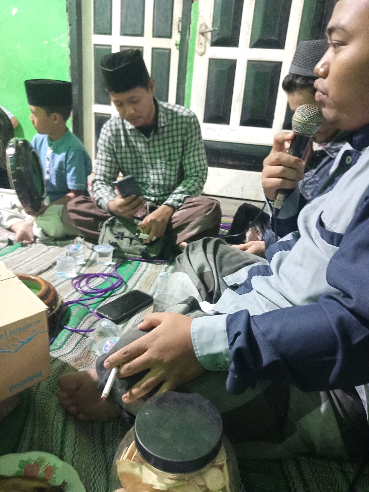 In a room with light green walls and a white door with square panels, four individuals are gathered on the floor, seated on a striped mat. The room appears to be in Ngablak, Indonesia, likely a residence.
In the foreground, a person is holding a lit cigarette, with smoke rising from it. Their right hand is visible, with fingers curved around the cigarette. This individual is wearing a light blue and dark blue striped shirt. To their left, another person is holding a microphone to their mouth, suggesting they might be speaking or singing. This person is wearing a dark grey peci, a traditional cap.
Further back, a man in a black peci is looking down at a mobile phone he is holding in his right hand. He is wearing a checkered shirt. Next to him, a young boy in a light blue shirt and a black peci is seated, facing to his left. A dark, circular object, possibly a musical instrument like a tambourine, is partially visible to his left.
Scattered on the mat are several items: two small plastic cups with clear liquid, a purple cord, a black mobile phone, a cardboard box with text and a blue bird logo, and a large glass jar containing what appears to be dried food items. The overall atmosphere suggests a casual gathering or informal event.