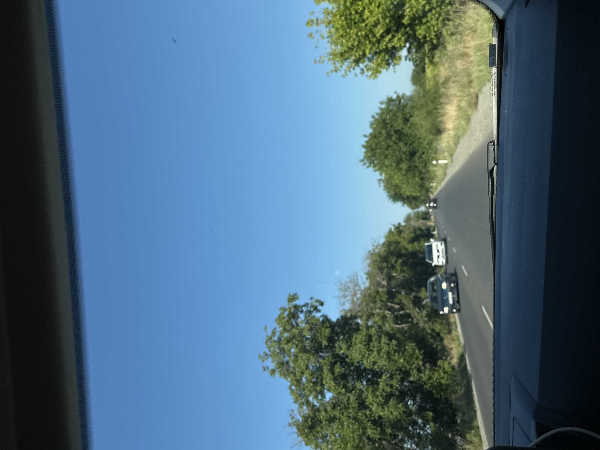 The image depicts a view from inside a vehicle looking out through the windshield. The perspective is from the driver's seat, showing the dashboard and the road ahead. It is a sunny day with a clear blue sky. The road is paved and appears to be a two-lane highway. There are several cars on the road, some in front and some in the distance, indicating traffic flow. Lush green trees line both sides of the road, with vegetation and grassy areas visible alongside. The scene suggests a drive through a rural or semi-rural area. Based on the location provided, this image was taken on a road in or near Kadievo, Bulgaria.