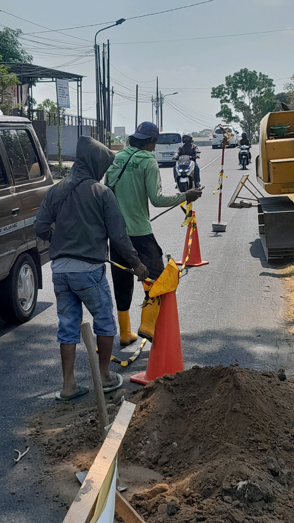In Tanjung, Indonesia, on a bright, sunny day, two construction workers are engaged in roadside maintenance. One worker, wearing a blue baseball cap, a light green long-sleeved shirt, and black pants, stands near an orange traffic cone, holding a yellow flag attached to a stick. The second worker, dressed in a dark grey hooded jacket, blue denim shorts, and flip-flops, is positioned closer to the camera, also holding a similar yellow flag on a stick. They are working on a paved road, with a pile of excavated dirt to their right. In the background, traffic passes by, including motorcycles and cars, and utility poles with numerous wires are visible. A parked brown vehicle is on the left side of the frame. The overall scene suggests ongoing roadwork in an urban or semi-urban setting.