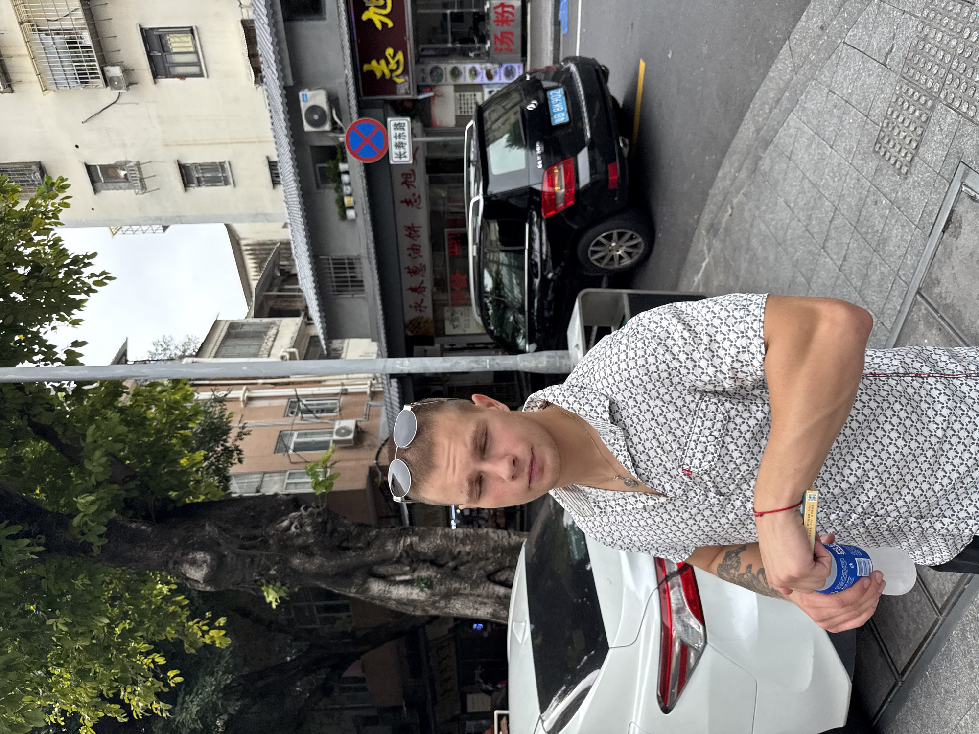 A young Caucasian man with short blonde hair and closed eyes stands on a sidewalk in Liwan District, China. He is wearing a short-sleeved patterned shirt and sunglasses perched on his head. His right hand is holding a blue bottle, and his left hand is holding a small box. He has a red string bracelet on his left wrist. To his left is the rear of a white car, and to his right is a black SUV. The background shows a street with buildings, trees, and street signs, including one with Chinese characters and a no parking symbol. The sky is overcast.