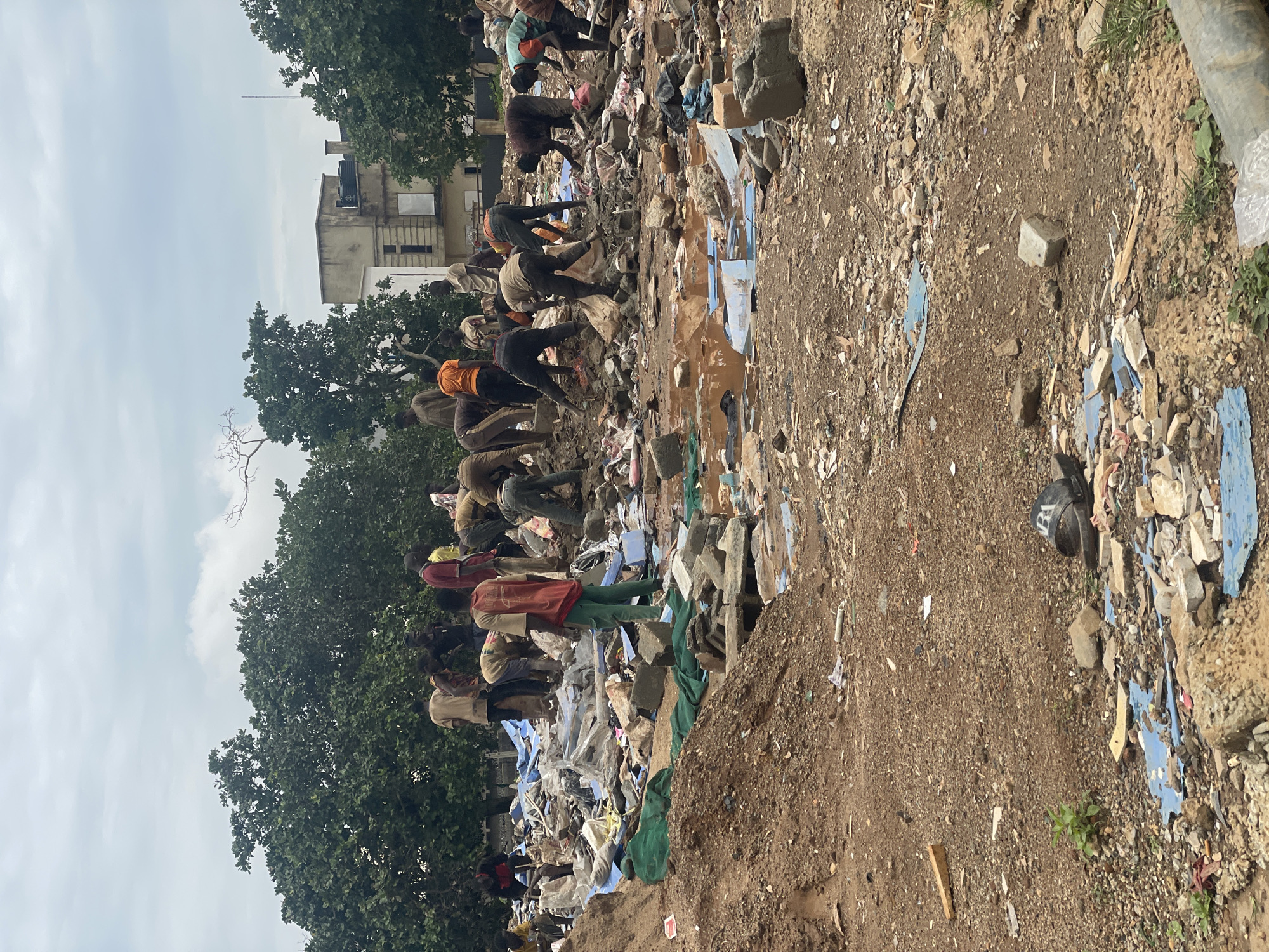 In Kurunduma, Nigeria, a group of approximately 20 individuals, appearing to be mostly young men and boys, are engaged in clearing debris from a large, open-air site. The ground is covered with a mixture of dirt, rubble, concrete fragments, and scattered blue tarps. The individuals are dressed in casual clothing, with some wearing brightly colored shirts. They are actively moving and sorting through the debris, some lifting heavy objects, others sifting through smaller pieces. The scene appears to be a manual labor effort to clean up a heavily littered area. In the background, a few trees and buildings are visible, suggesting an urban or semi-urban environment. The sky is overcast.