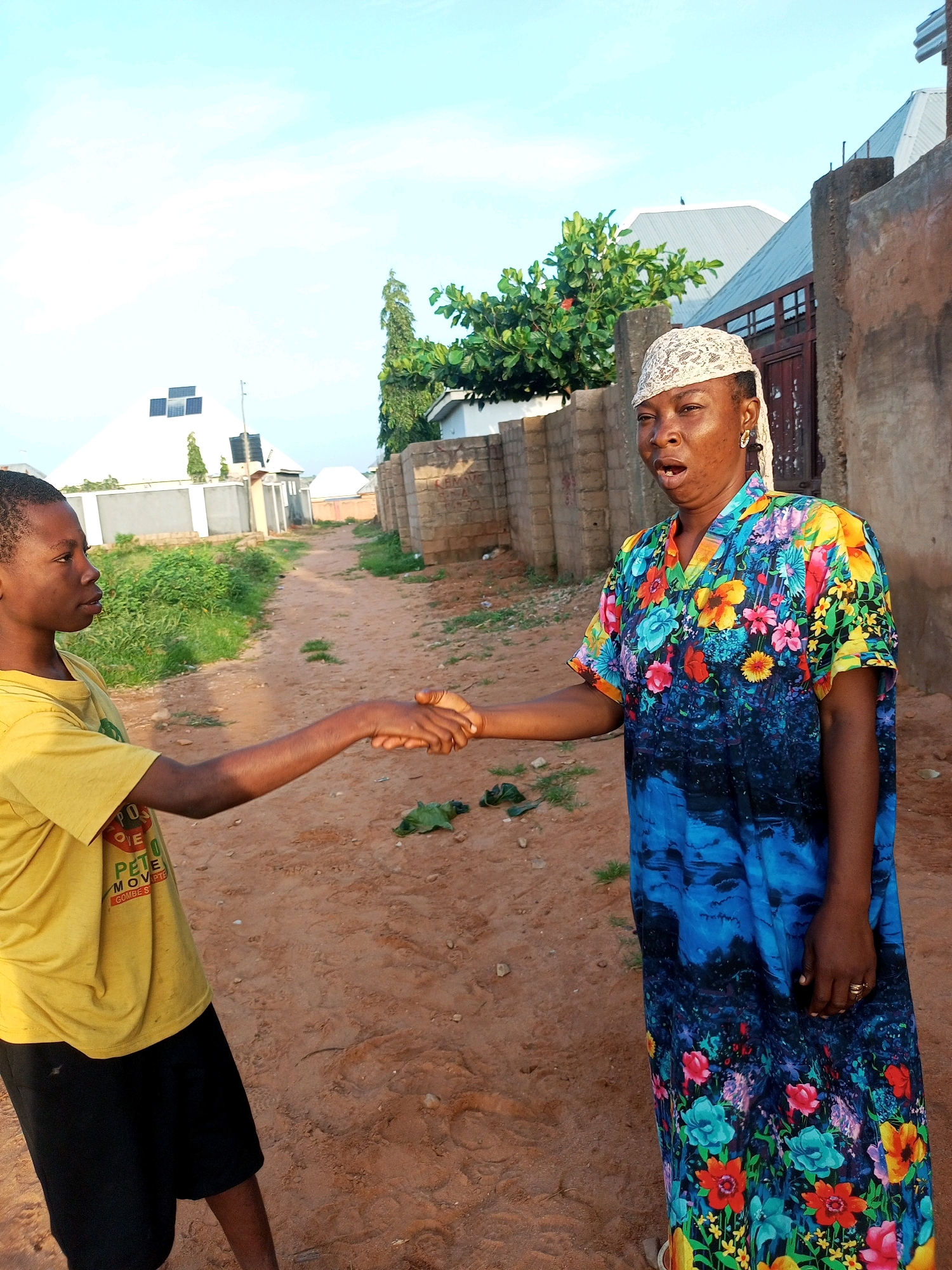The image captures a moment in Tempure, Nigeria, where two individuals are engaged in a handshake. The background reveals a semi-urban environment with a dirt path leading into the distance. On the left, a young person wearing a yellow t-shirt and black shorts stands facing right, their arm extended for the handshake. On the right, an adult is dressed in a vibrant, floral-patterned blue dress and a white head covering. Their mouth is open, suggesting they are speaking. Buildings constructed with concrete blocks and a white-colored building with solar panels are visible in the distance, bordering the path. Green vegetation lines the sides of the path, and the sky above is a pale blue with some cloud cover. The overall scene depicts a casual interaction on a dusty street.