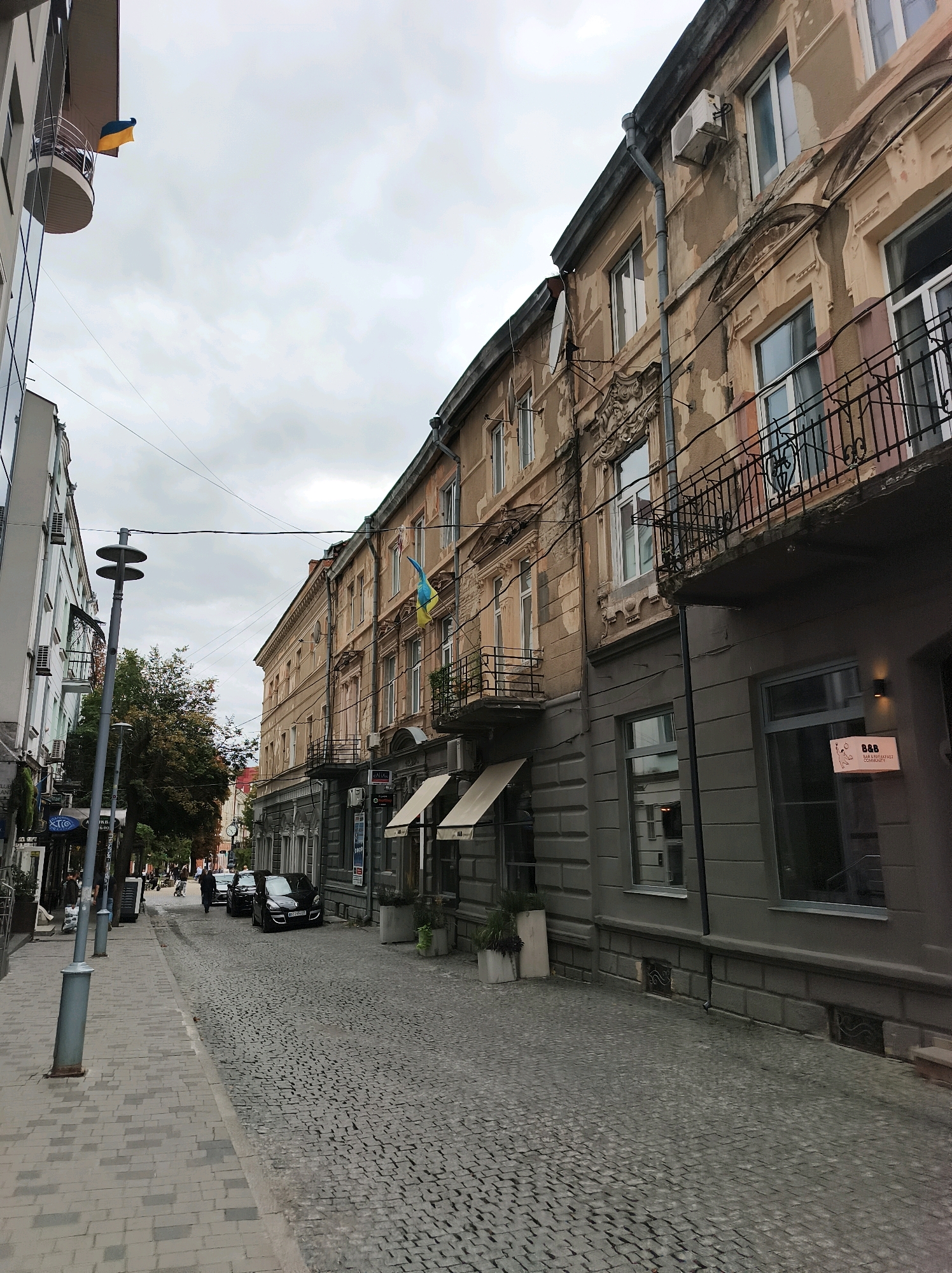 The image depicts a street scene in Ternopil, Ukraine. The street is paved with cobblestones, and lined with buildings on both sides. On the left, a modern building with glass windows and a balcony is visible. On the right, an older building with weathered stucco and balconies with wrought-iron railings stands. A Ukrainian flag is displayed on the older building. The sky is overcast. There are several cars parked on the street and a lamppost on the left side. People are walking on the sidewalk on the left. The overall scene suggests a street in a city with a mix of old and new architecture.
